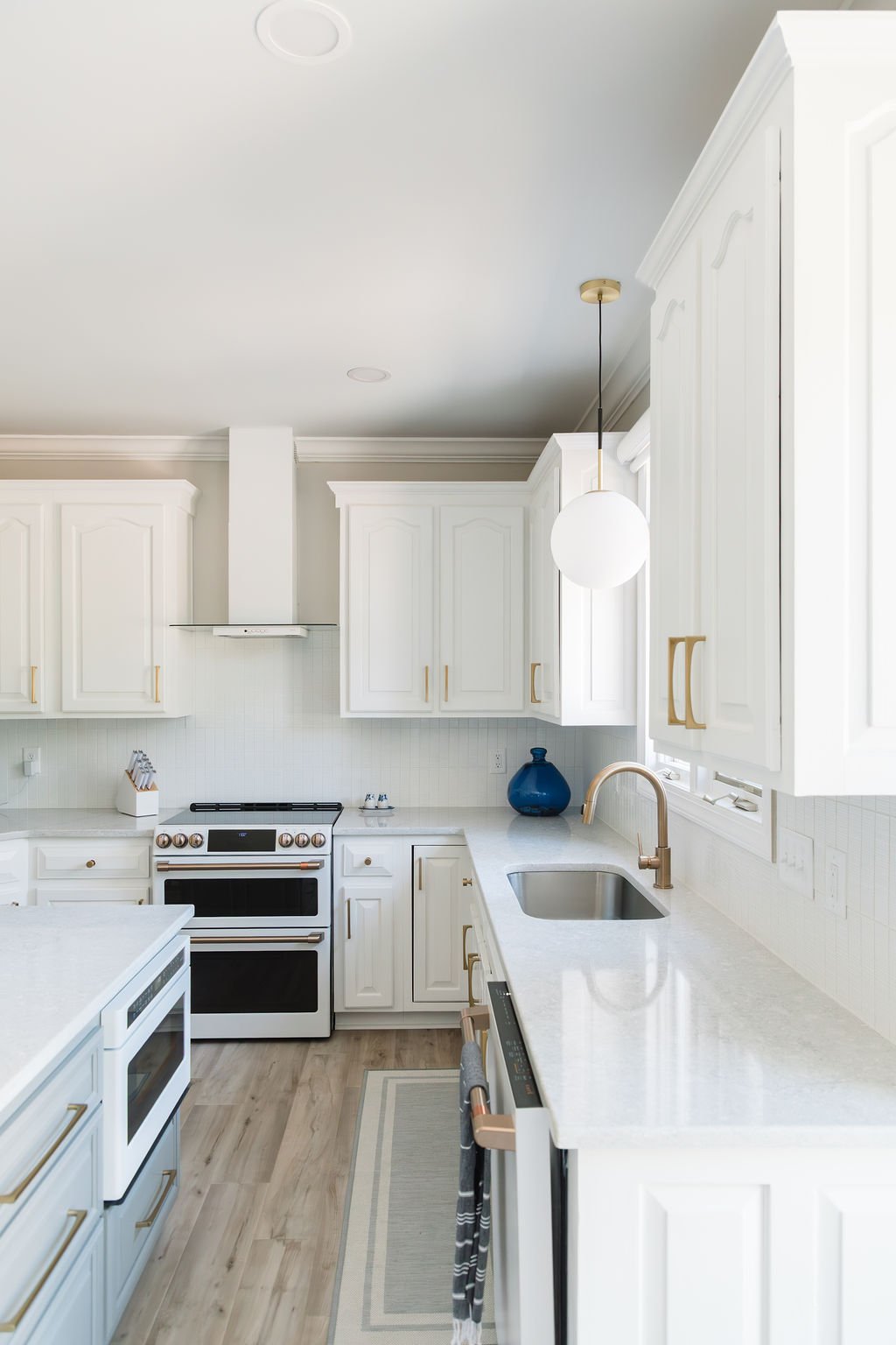 Bright white kitchen with gold accents, white cabinets, white countertops, stainless steel sink, black and white oven, blue vase, and hanging globe pendant light.