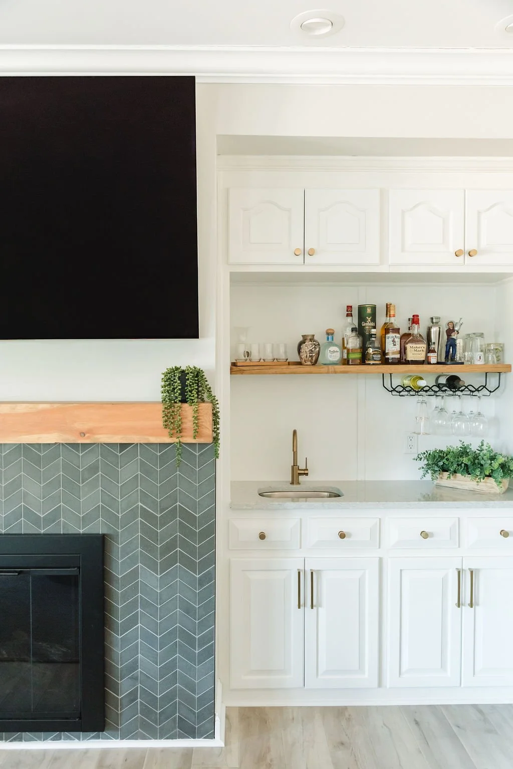 Part of a kitchen with white cabinets, a small sink, and a wooden shelf holding liquor bottles and glasses. A black chalkboard, a greenery plant, and a fireplace with a chevron tile pattern are also visible.