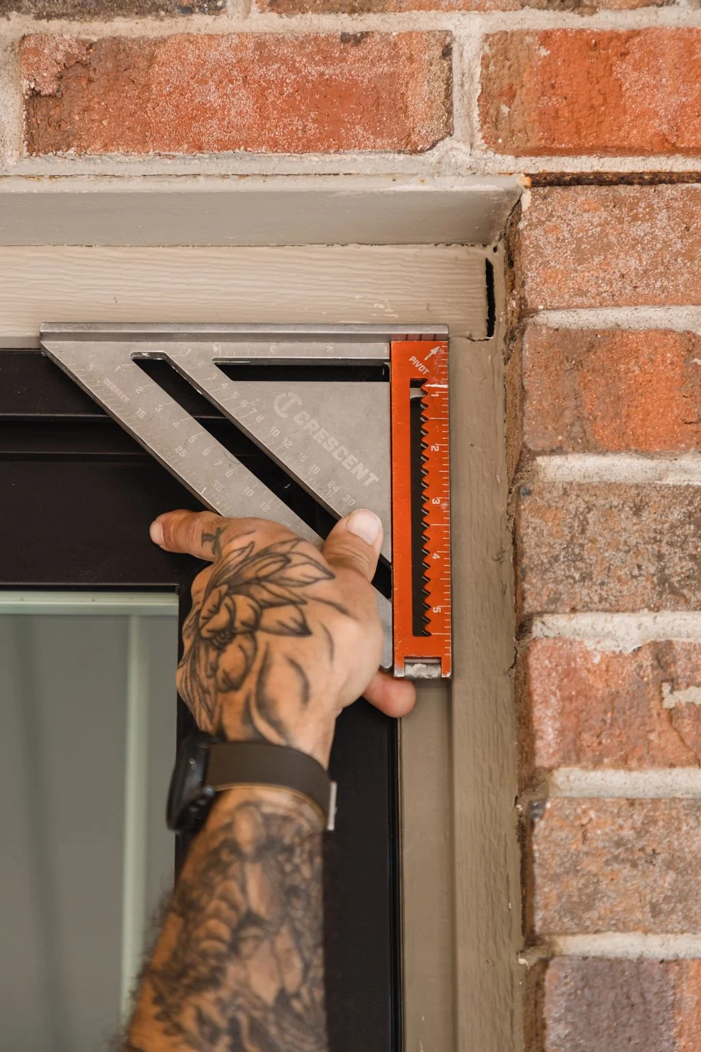 A person with tattooed arms measuring the height of a door frame with a carpenter's square and a pencil, near a brick wall.
