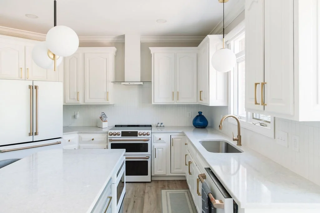 Bright white kitchen with gold handles, white cabinets, and a white countertop. Features a stove, refrigerator, and a sink under a window, with pendant lighting fixtures.