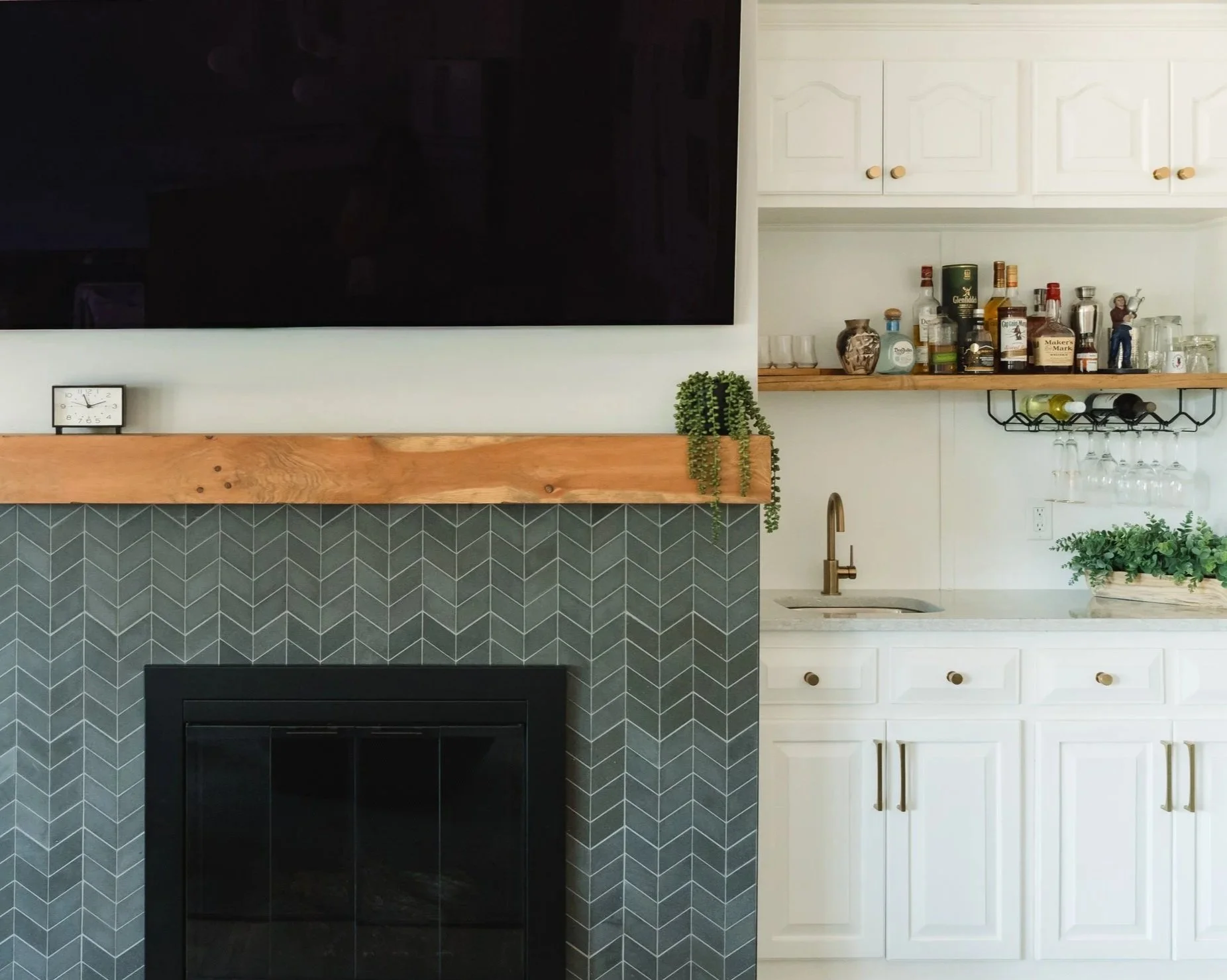 Living room with a fireplace, a mounted flat-screen TV, a small clock, decorative plants, and a kitchen area with white cabinets, a wooden shelf with bottles and glasses, and a sink.
