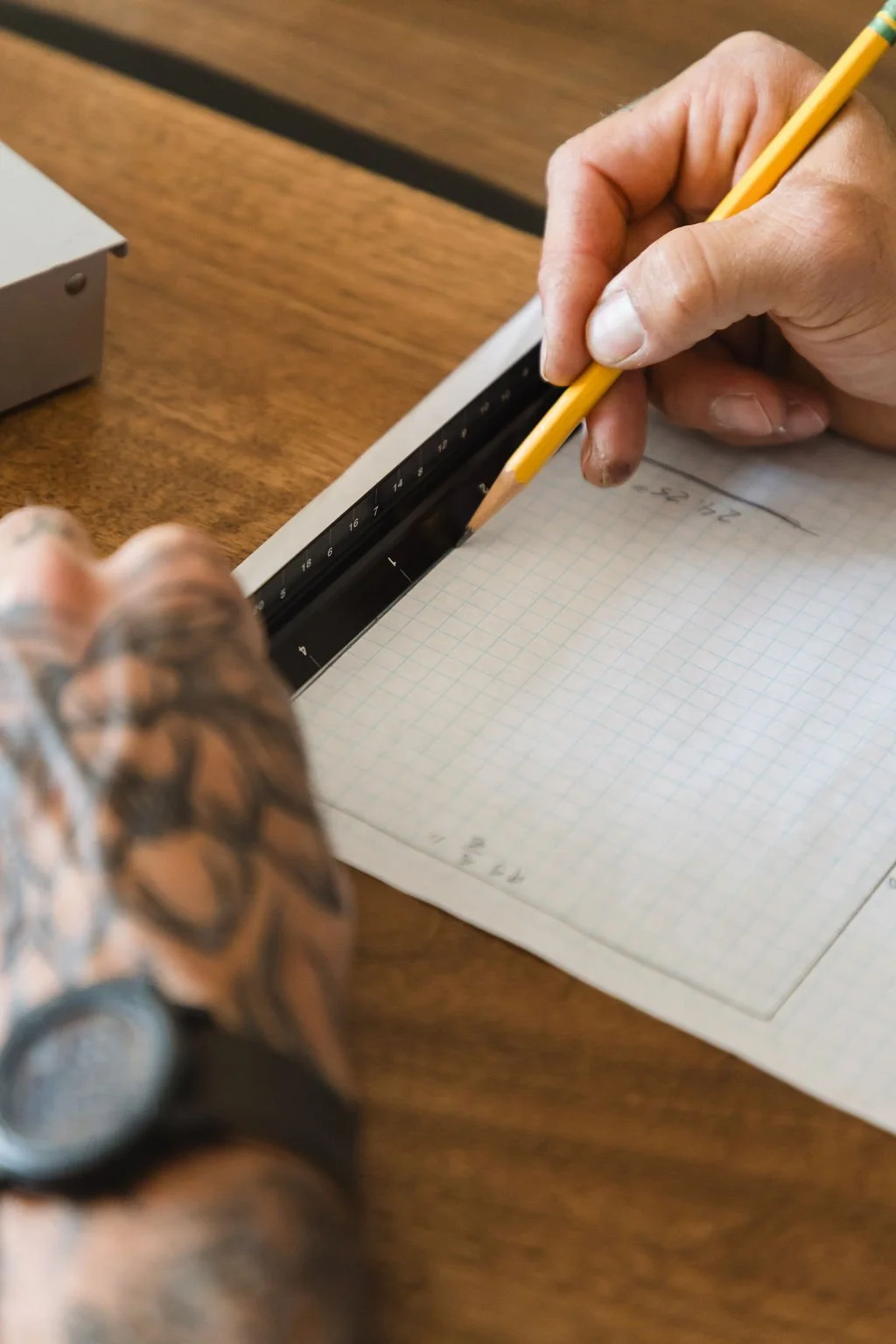 A person using a yellow pencil to draw a line on graph paper with a metal ruler, on a wooden desk.