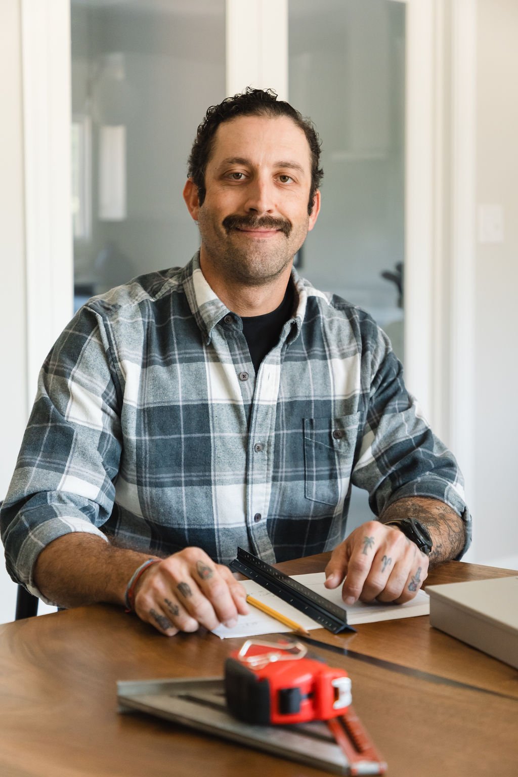 Jay Southers wearing a plaid shirt, is sitting at a wooden table with a measuring tape, ruler, pencil, and notepad, smiling at the camera.