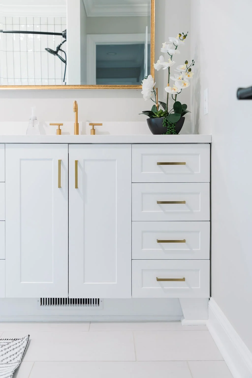 A modern bathroom vanity with white cabinetry, gold handles, and a white countertop. A black vase with white orchids sits on the right side of the vanity. A mirror with a gold frame is above the vanity, and a gold faucet is centered. Part of a shower with white tiles is visible in the reflection.