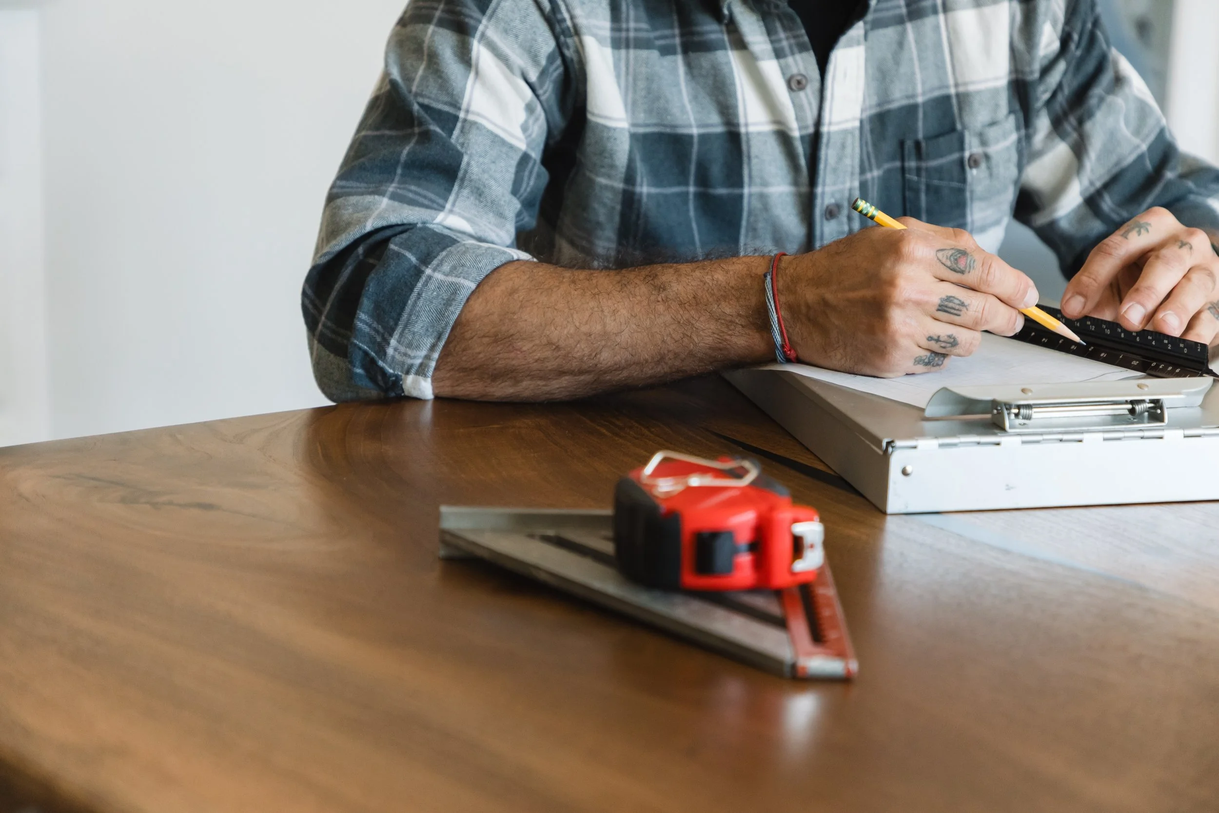 Person with tattoos on hands and tattoos on fingers wearing a plaid shirt working with a ruler and pencil on a wooden table.