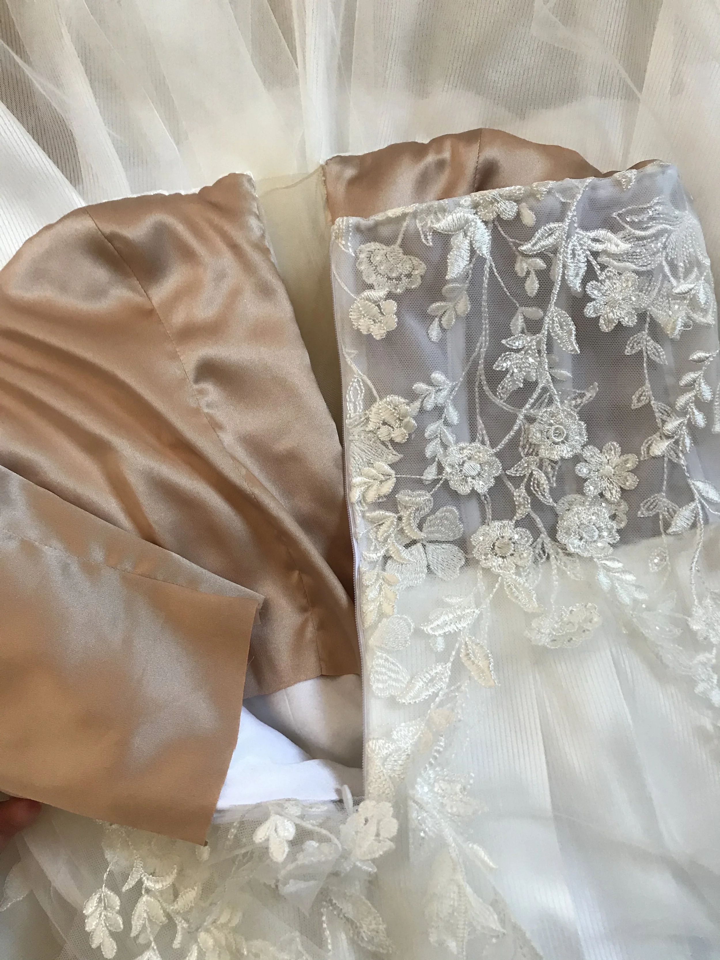 Nahaufnahme eines Brautkleides mit weißen Spitzenapplikationen und cremefarbenem Satin. Das Kleid hat eine transparente Schulterpartie mit floralem Stickmuster.