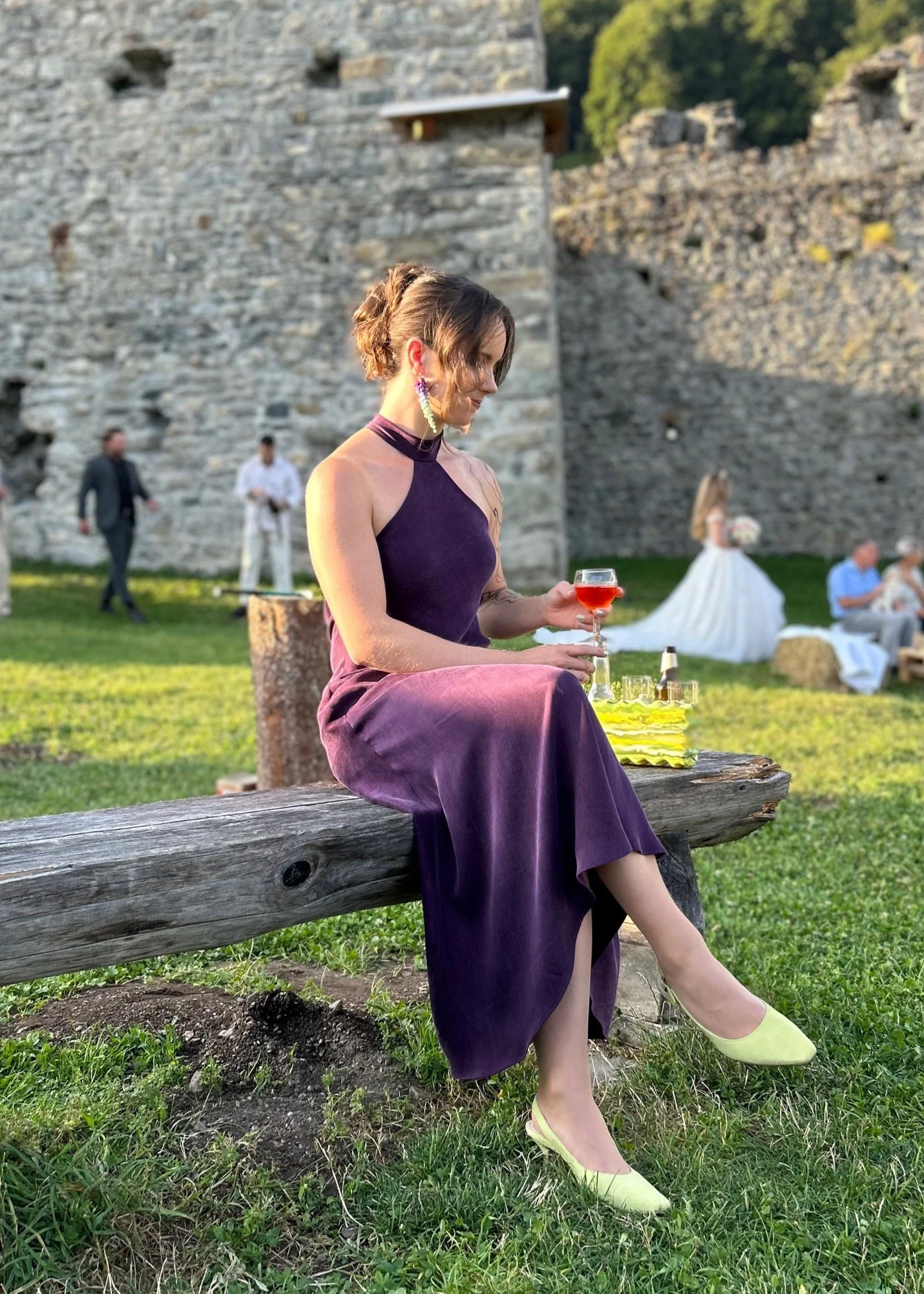Eine Frau in einem lila halterneck Kleid sitzt auf einer Holzbank im Freien und hält ein Glas mit Aperol. Im Hintergrund sind mehrere Personen, eine Braut im weißen Kleid und andere in formeller Kleidung.