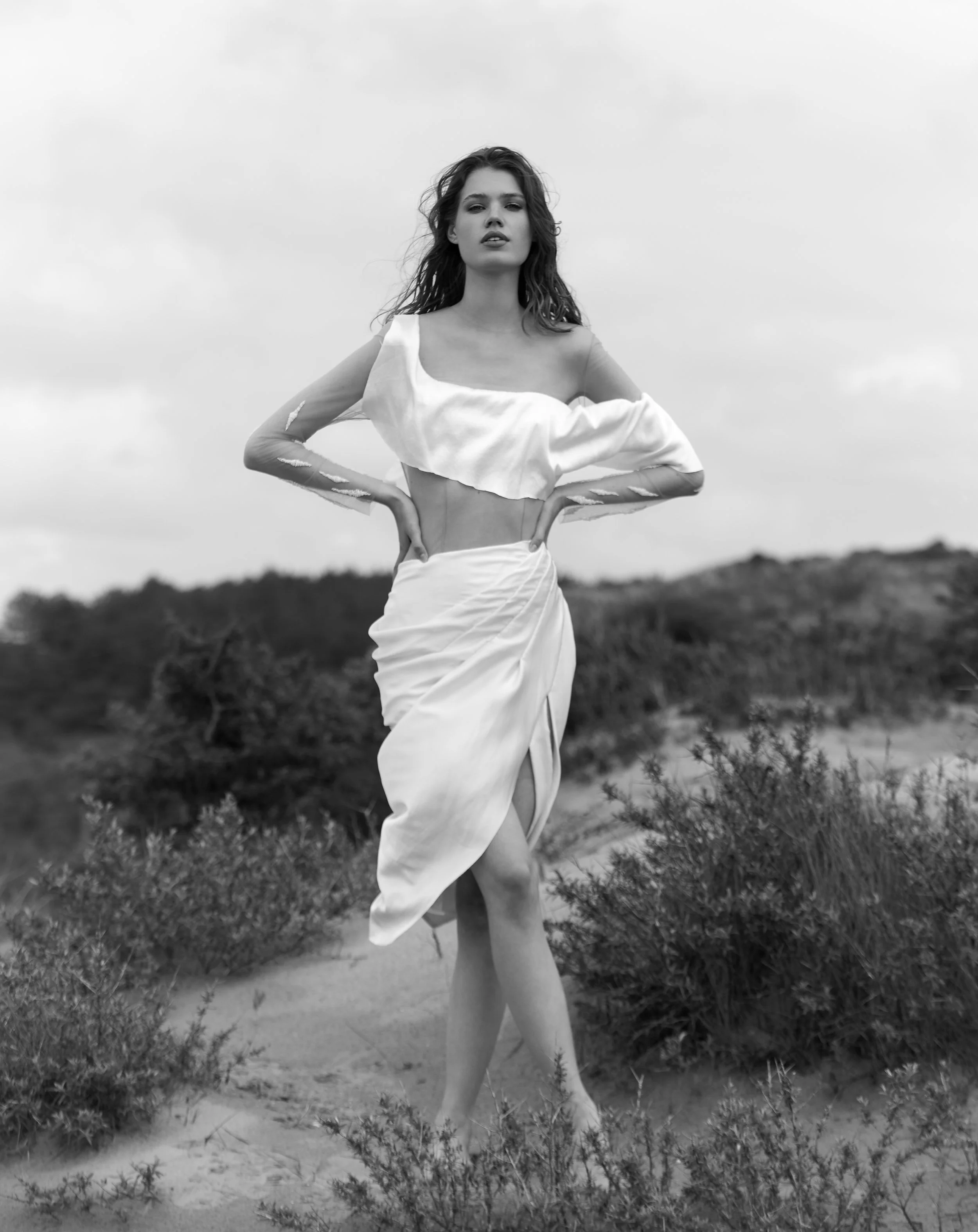 Schwarz-Weiß-Bild einer Frau, die in einem Off-Shoulder-Kleid in einer natürlichen Umgebung steht, mit einem bewölkten Himmel im Hintergrund.