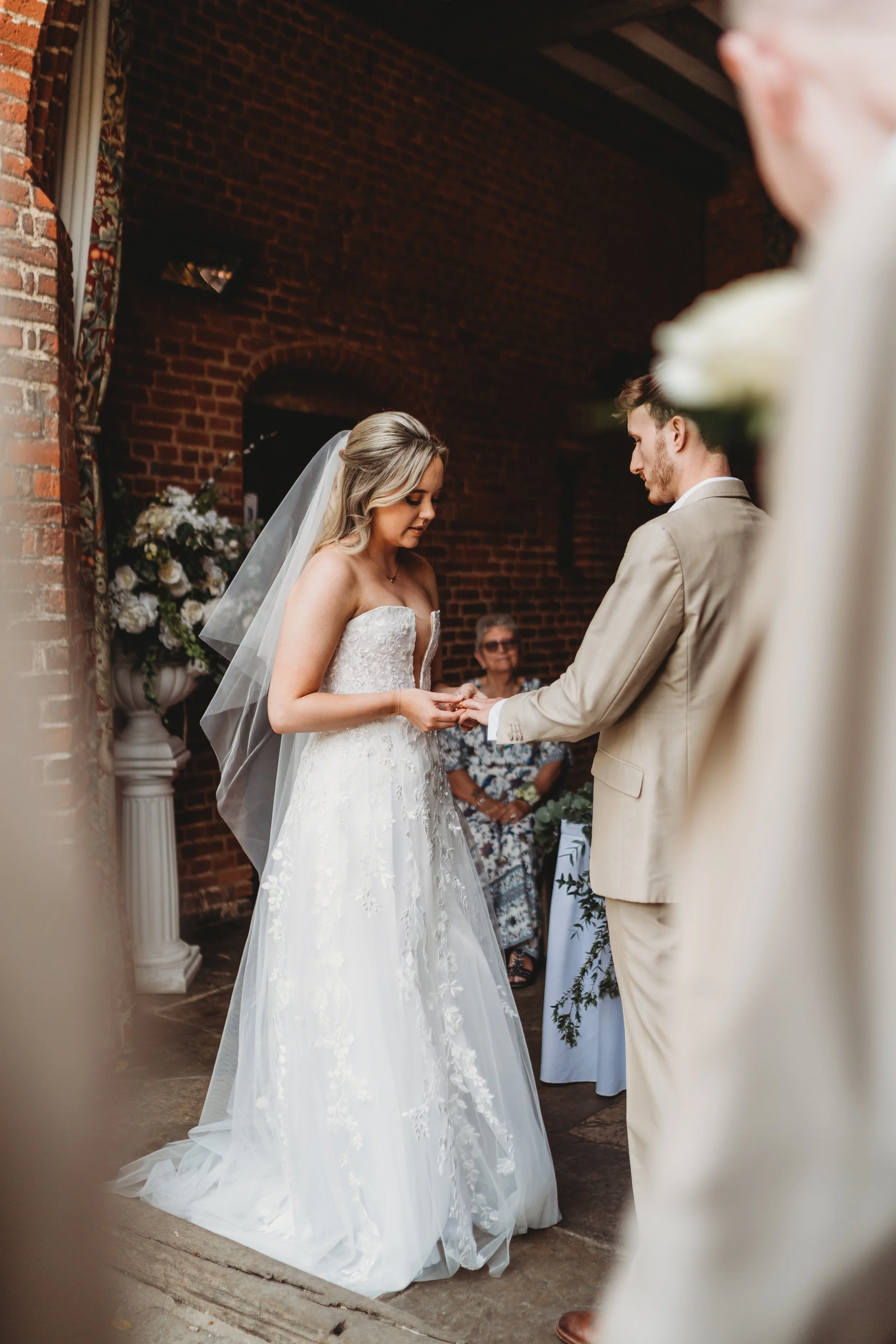 Ein Brautpaar bei einer Eheschließung, die Frau trägt ein weißes Hochzeitskleid mit Schleier und der Mann einen beige Anzug, im Hintergrund sind rote Backsteinwände und eine Person, die einen Blumenstrauß hält.