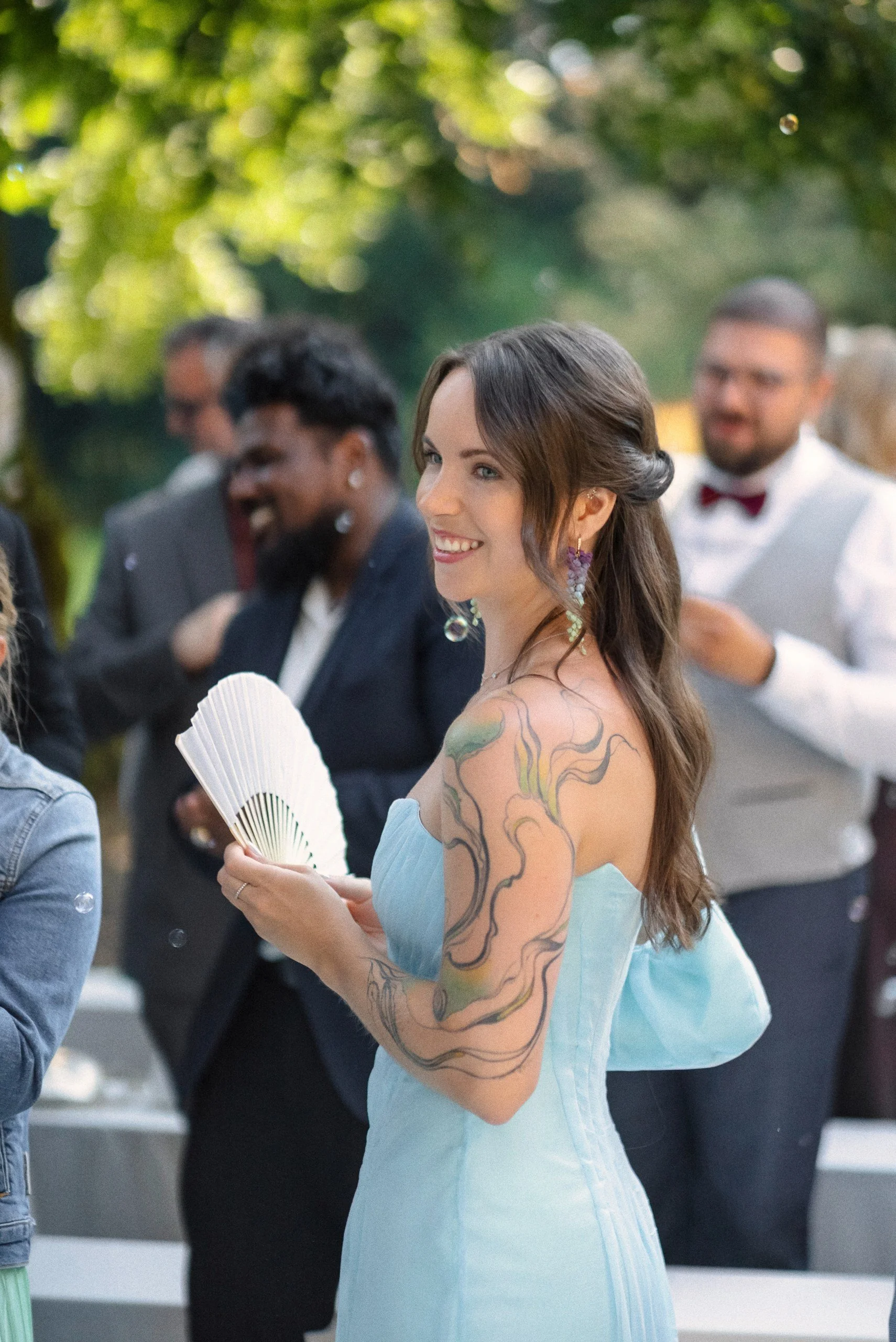 Eine junge Frau trägt ein hellblaues Kleid, hält einen Fächer und hat bunte Tattoos auf ihrem Arm. Sie lächelt und ist umgeben von Menschen, die im Freien bei Tageslicht feiern.