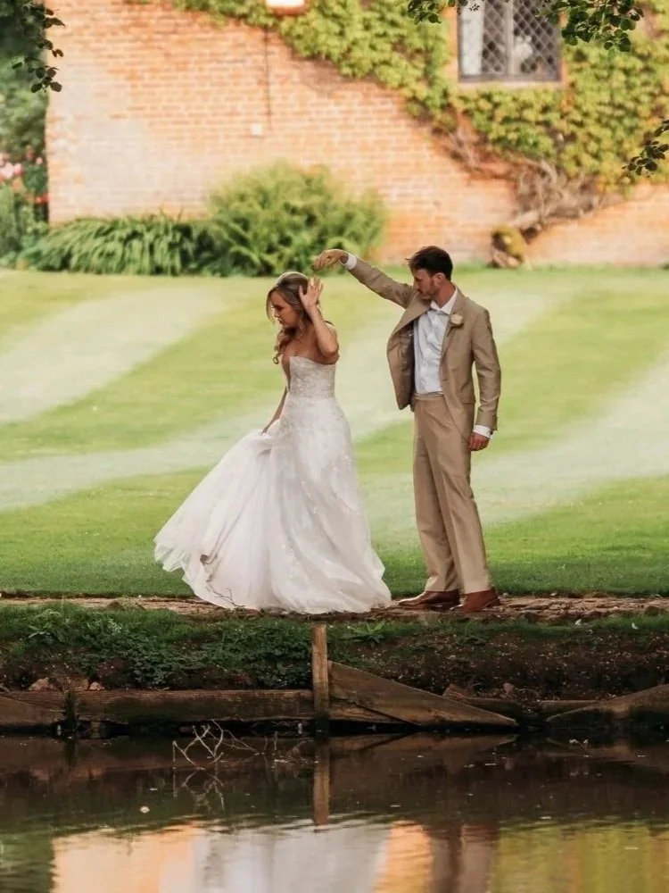 Ein Brautpaar tanzt am Flussufer im Freien, die Hochzeit befindet sich im Sommer, umgeben von Bäumen und einem alten Backstein-Gebäude im Hintergrund.