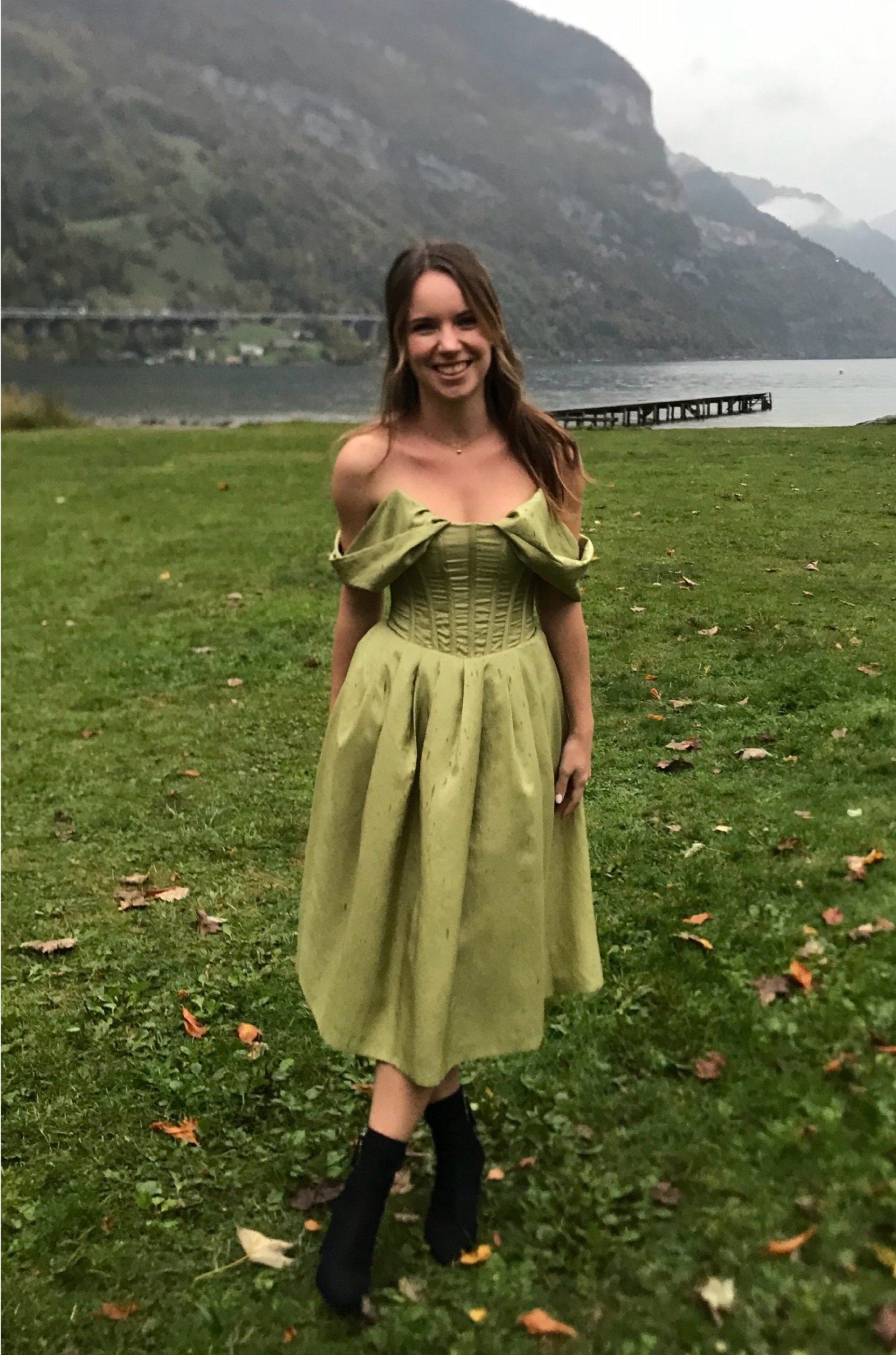 Junge Frau in einem grünen Kleid steht auf einer Wiese mit einem See und Bergen im Hintergrund.