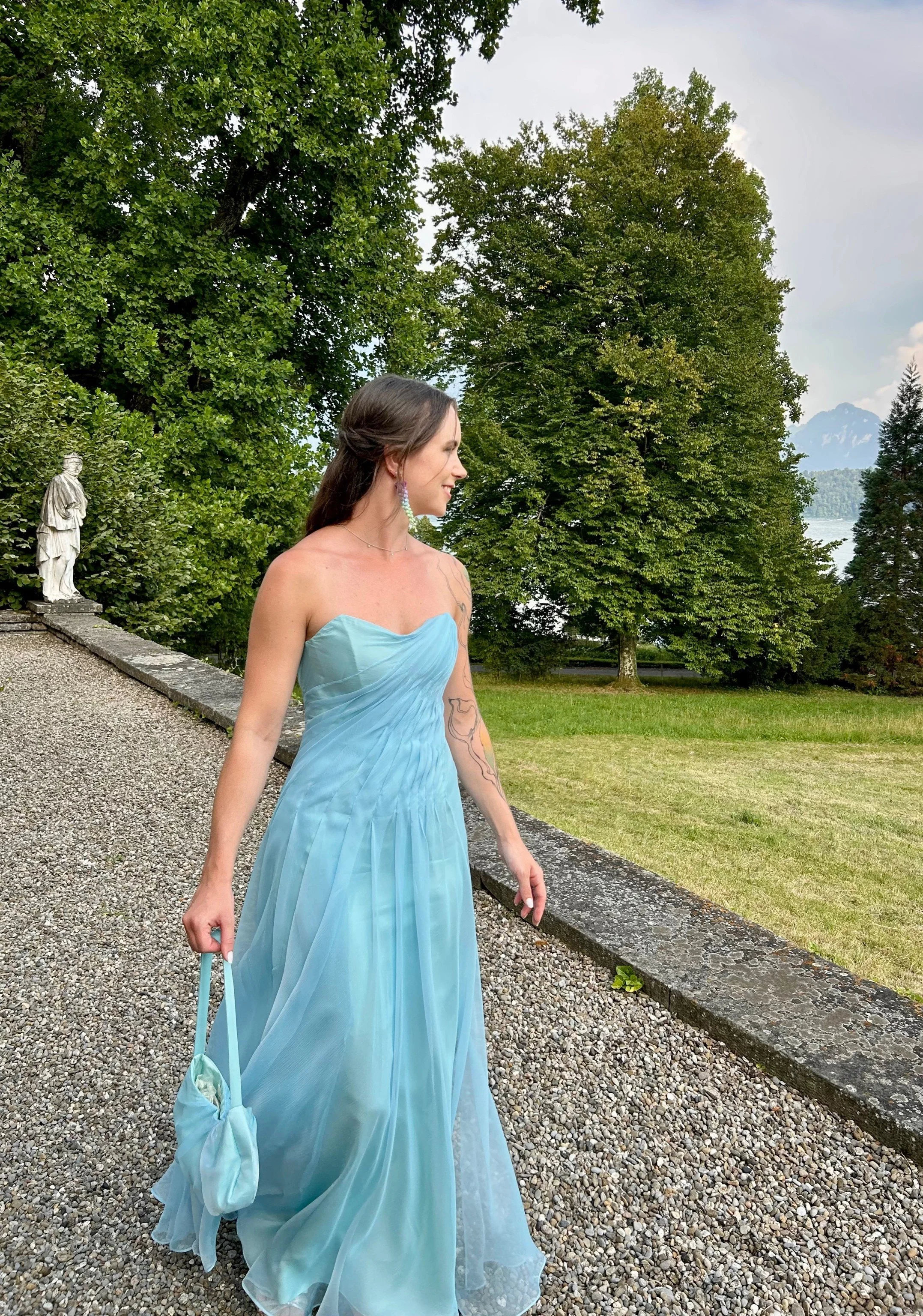 Eine Frau in einem eleganten, hellblauen Abendkleid  läuft an einer Mauer entlang. Im Hintergrund sind grüne Bäume, ein See und Berge zu sehen.