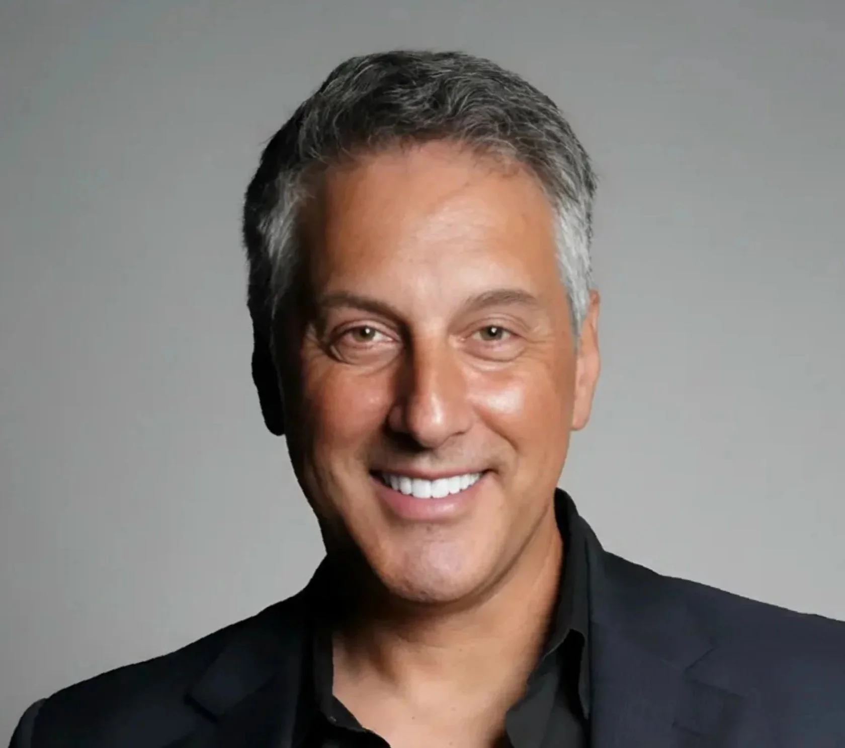 A smiling man whose name is Mike Goos with gray hair wearing a black shirt against a neutral gray background.