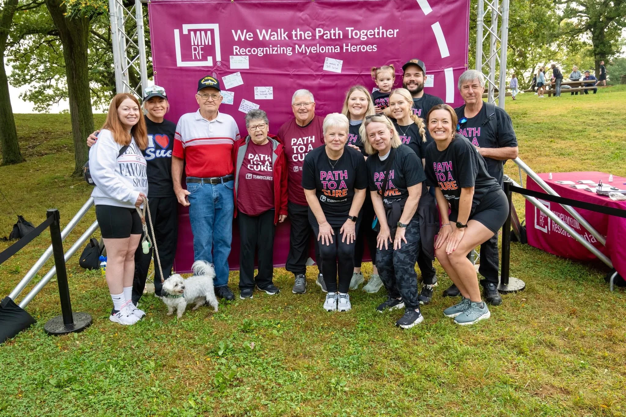Group of people posing outdoors in front of a purple banner that reads, 'We Walk the Path Together, Recognizing Myeloma Heroes.' The group includes men, women, children, and a small dog, and appears to be participating in a charity walk event.