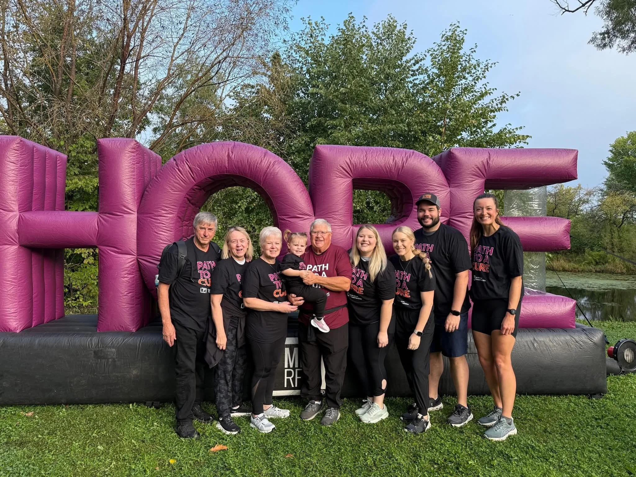A group of people posing in front of a large pink inflatable sculpture outdoors during daytime. The group includes both children and adults, all smiling.