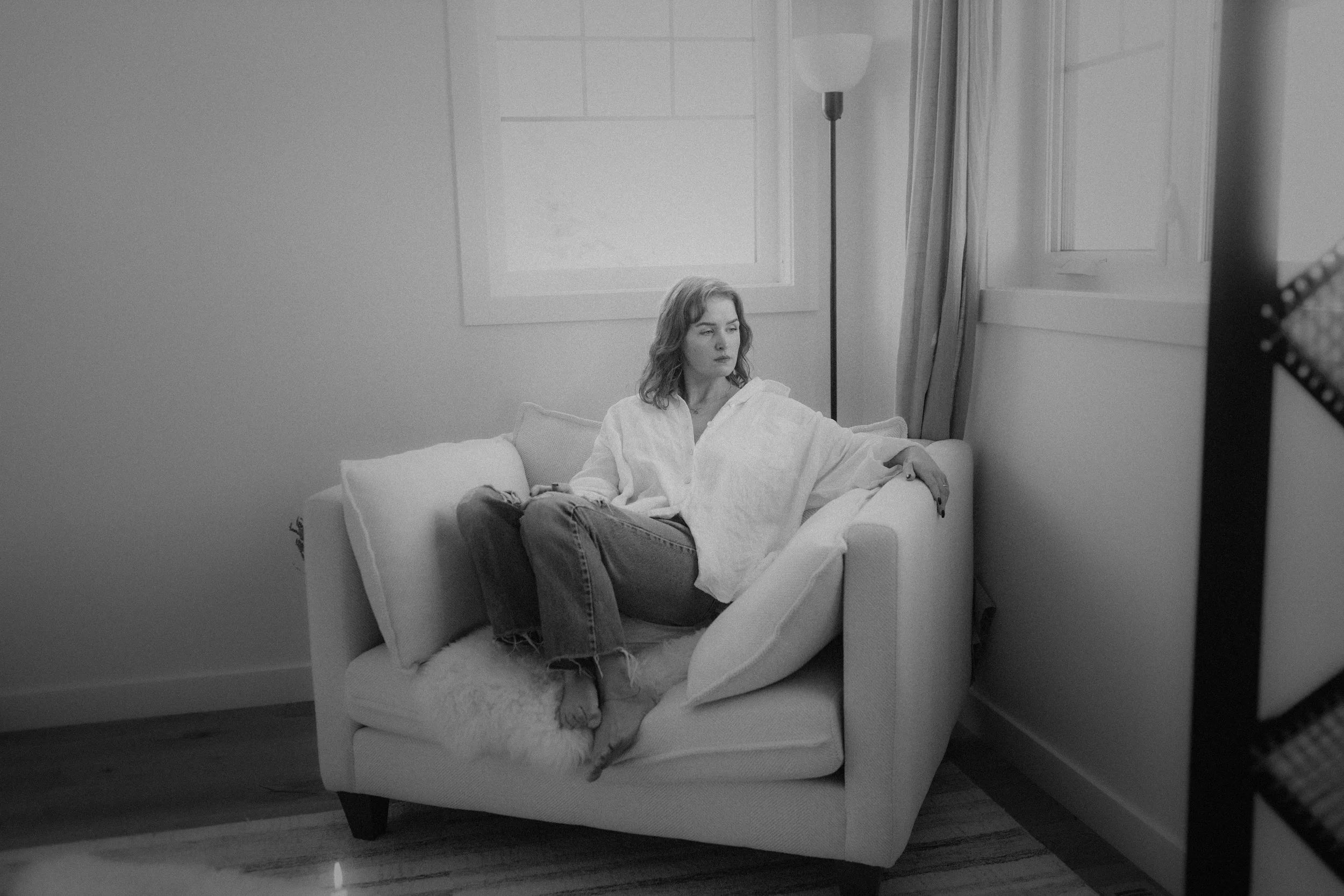 A woman sitting on a sofa by the window in a room with a lamp and curtains.