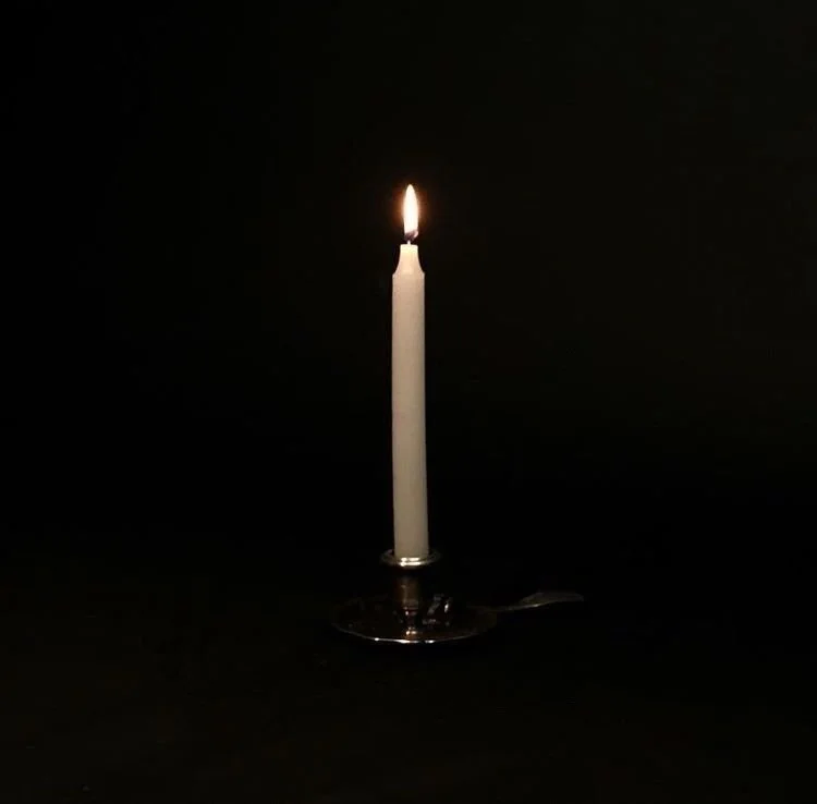 A single white candle burning on a metallic holder in a dark room