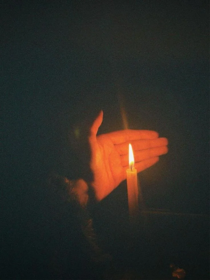 A person holding a lit candle with both hands in a dark environment, casting a warm glow.