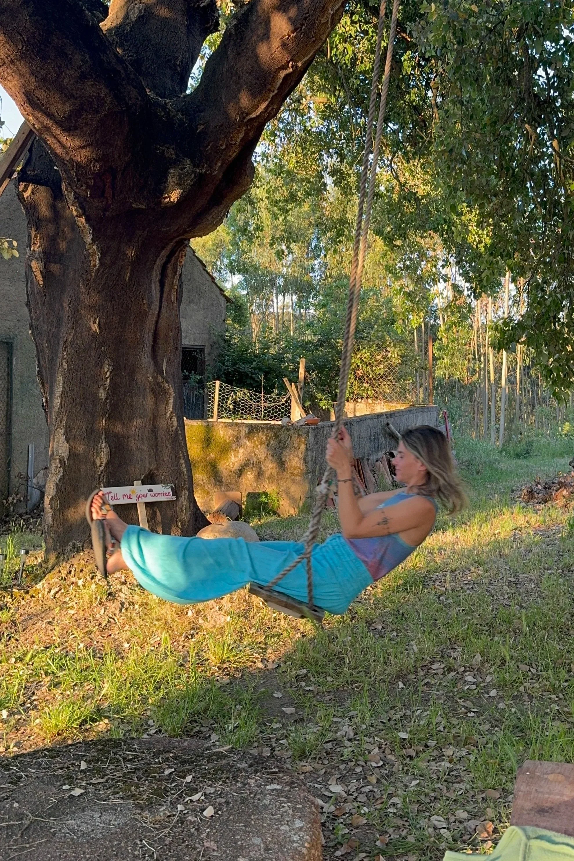 Woman sitting on a swing tied to a large tree outside during sunset, holding the rope with her right hand and dressed in a sleeveless top and turquoise pants.