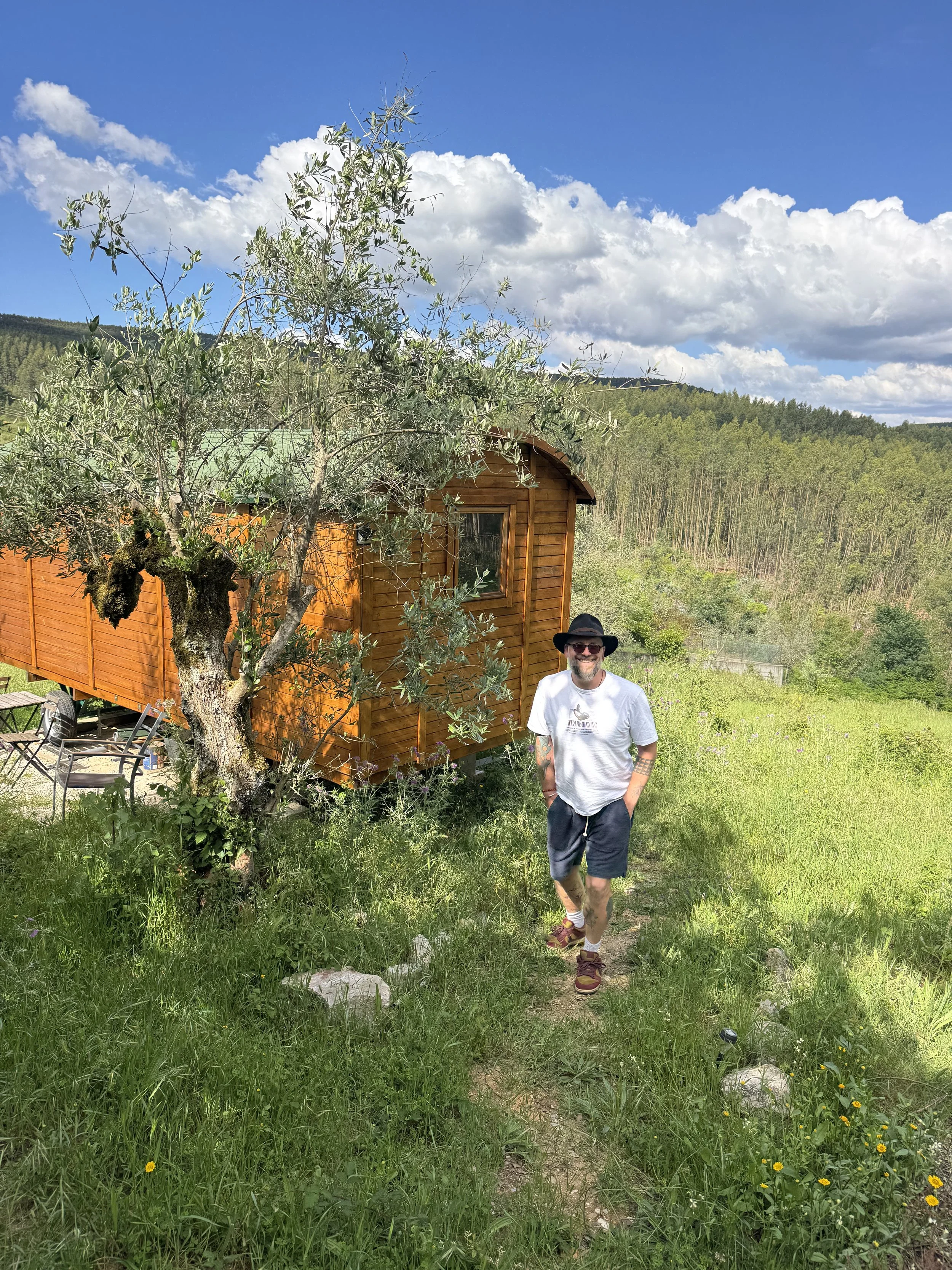 A man with sunglasses, a hat, a white t-shirt, and shorts, standing on a grassy path outdoors, with a wooden tiny house, a tree, and a forested mountain in the background under a partly cloudy sky.