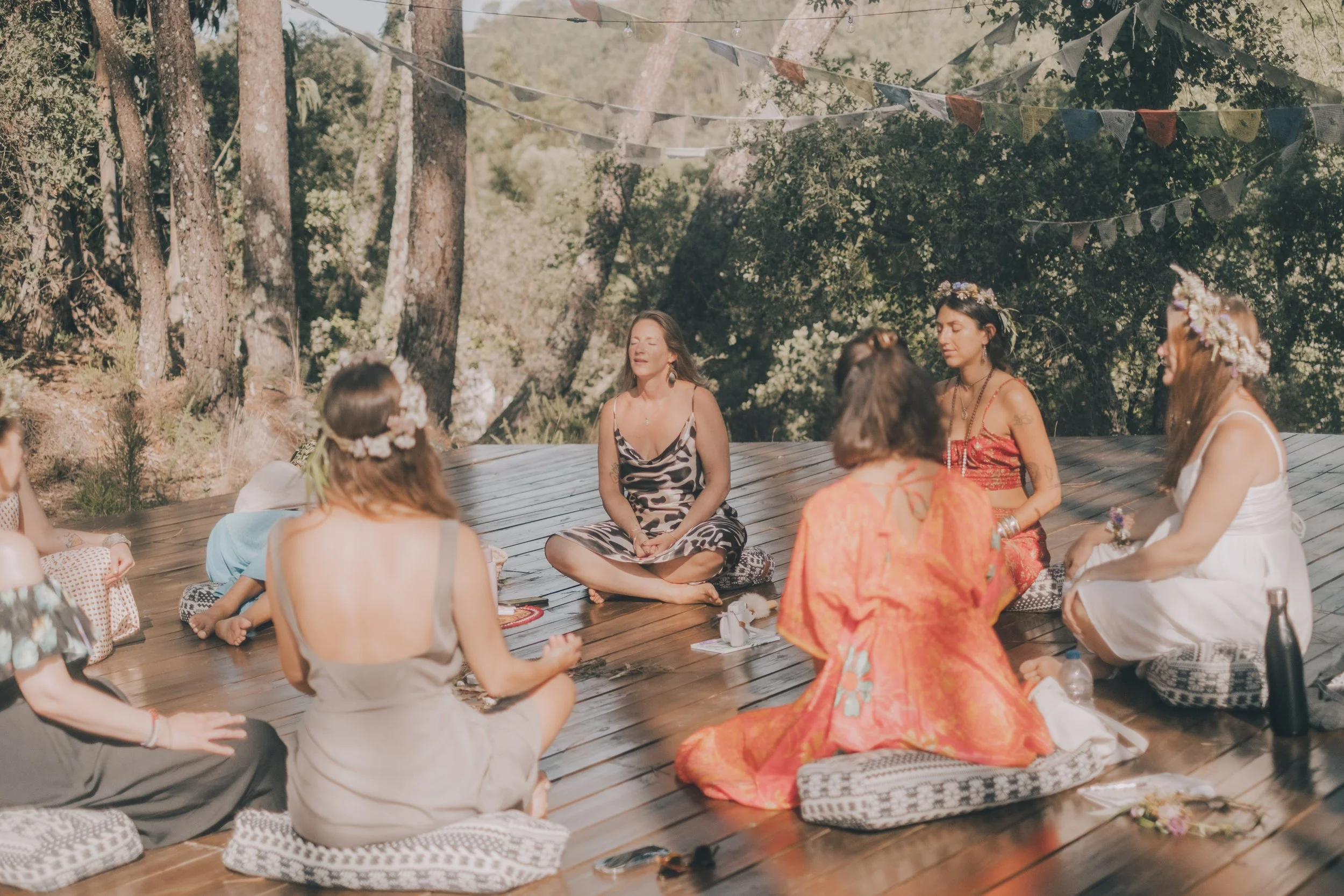 A group of women sitting in a circle on wooden floorboards outdoors during daytime, participating in a meditation or yoga session. They are surrounded by trees and decorated with string lights and colorful flags.