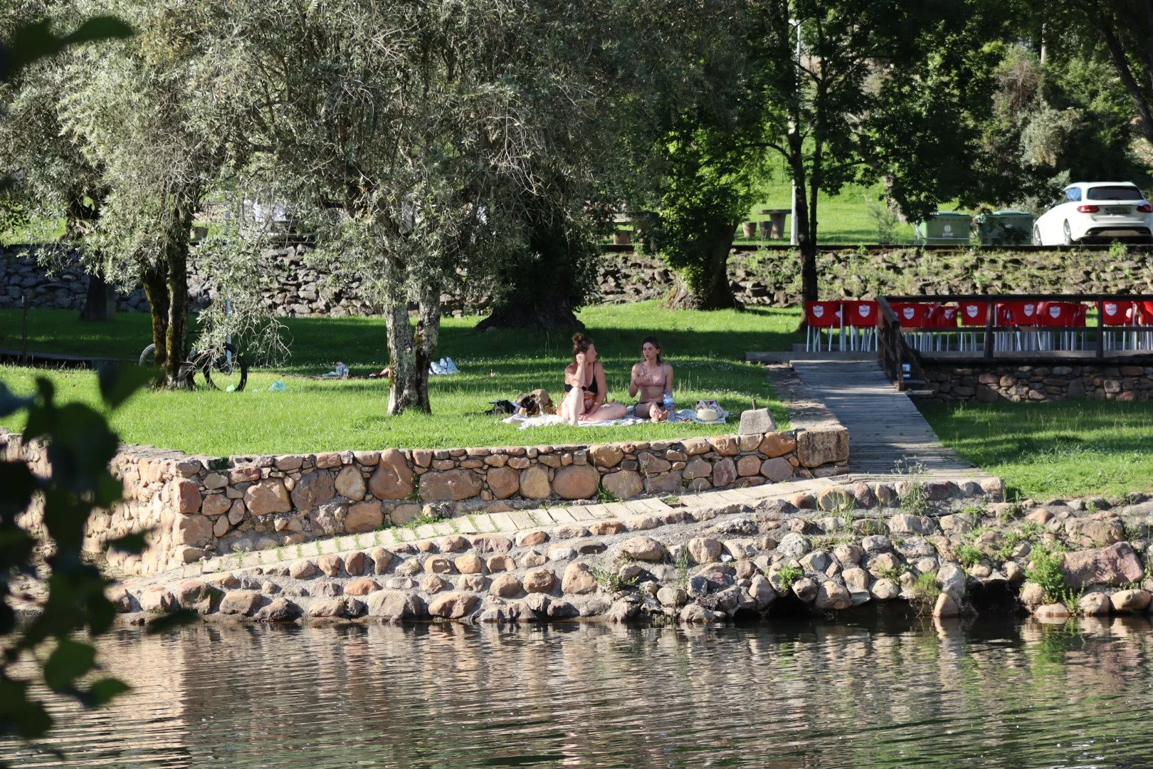 Two women sitting on a blanket by a river, surrounded by trees and a stone wall, with chairs and cars in the background.