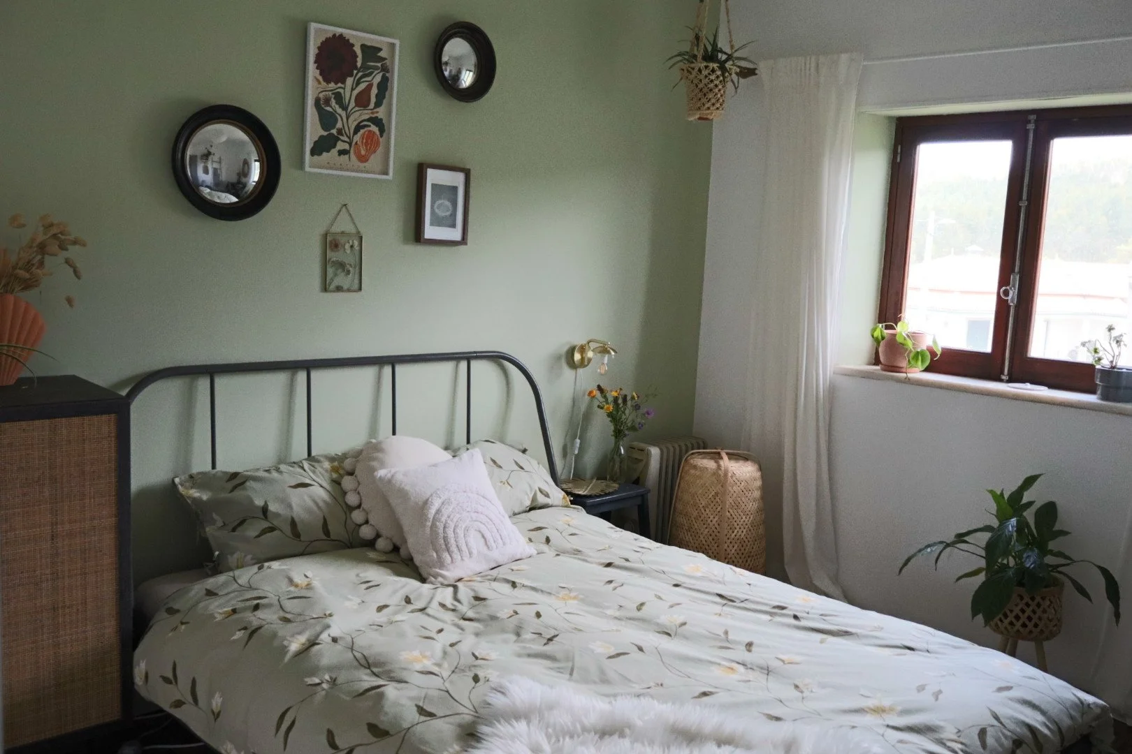 A cozy bedroom with a black metal bed frame, floral bedding, and white decorative pillows. The green wall above the bed features framed artwork and mirrors. A bedside table holds a vase with flowers, and a window with white curtains lets in natural light. Potted plants are on the windowsill and nearby surface.