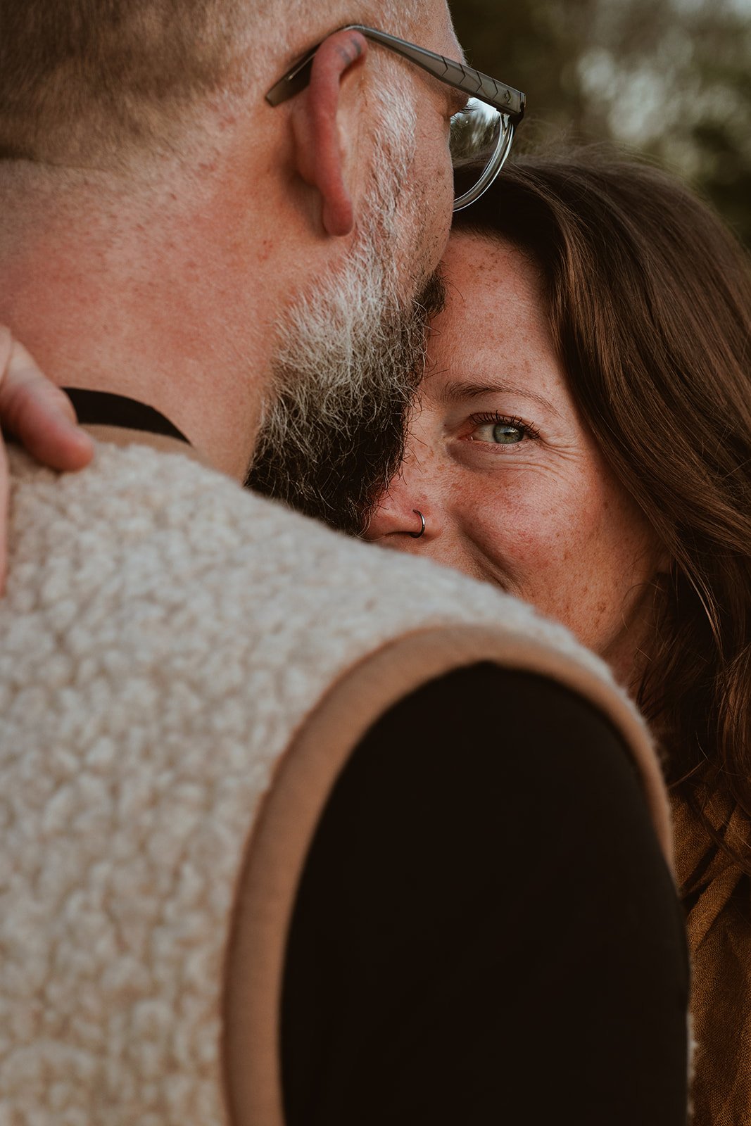 A close-up of a man and woman embracing, with the man’s face partially visible and the woman smiling. The woman has a nose piercing and freckles.