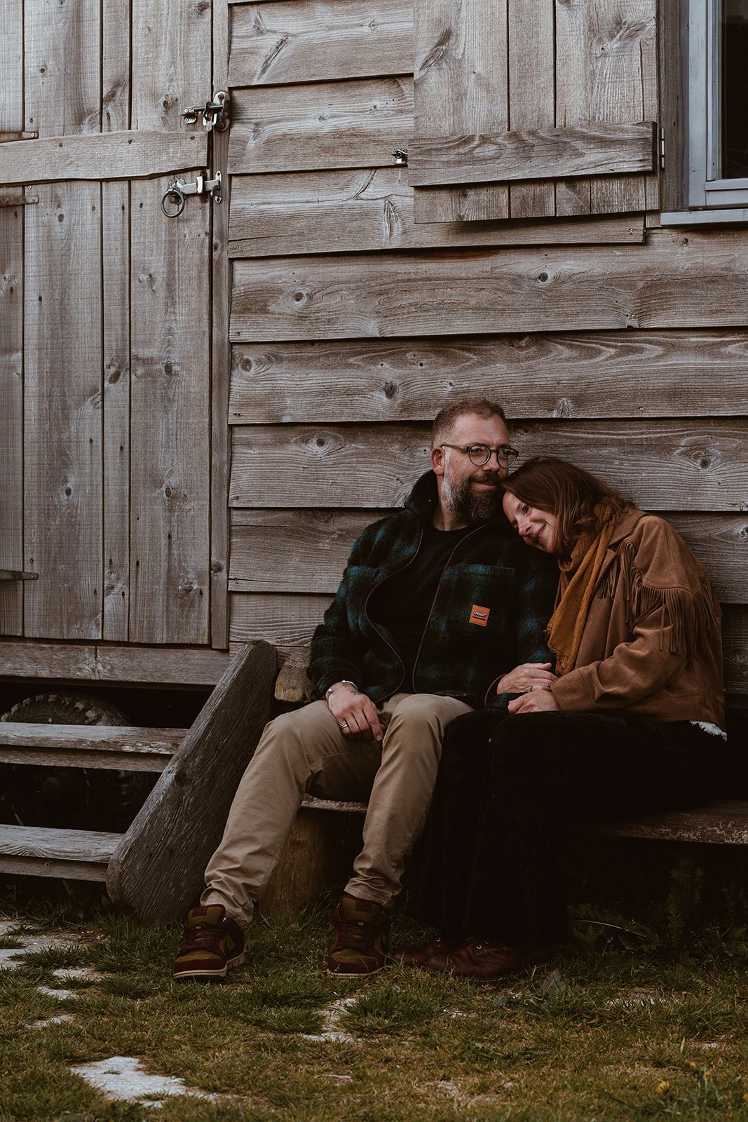 A man and a woman sitting together on a wooden bench, leaning their heads against each other, outside a rustic wooden building.