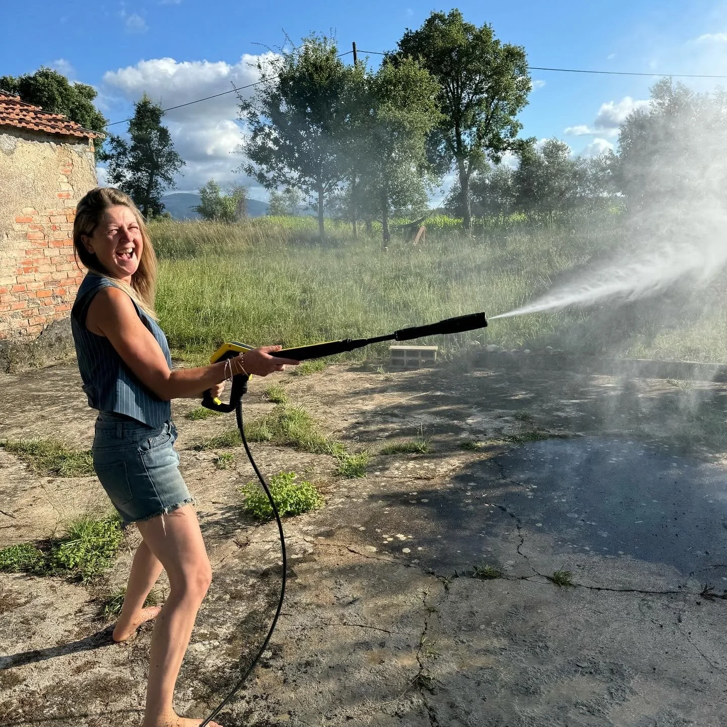Woman using a pressure washer outdoors, laughing, with trees, grass, and a brick building in the background on a sunny day.