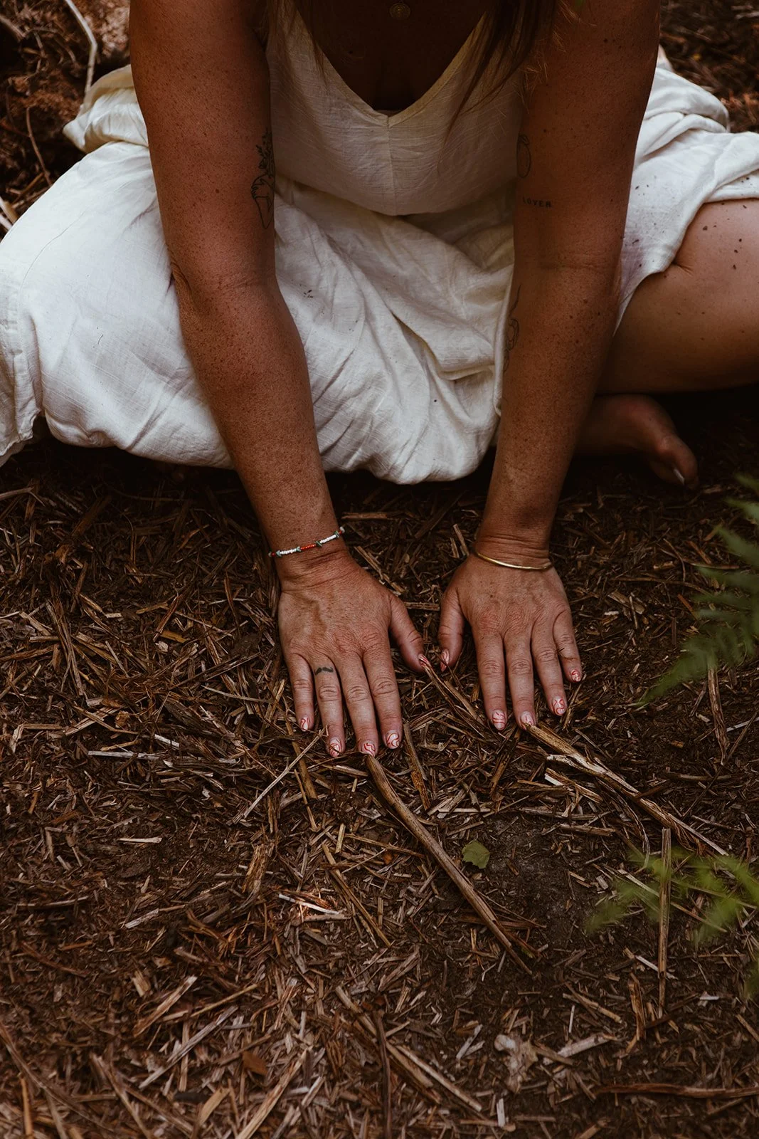 A person kneeling on soil with hands on the ground, wearing a light-colored dress, with tattoos on their arms and hands, and jewelry including a bracelet and a bangle.