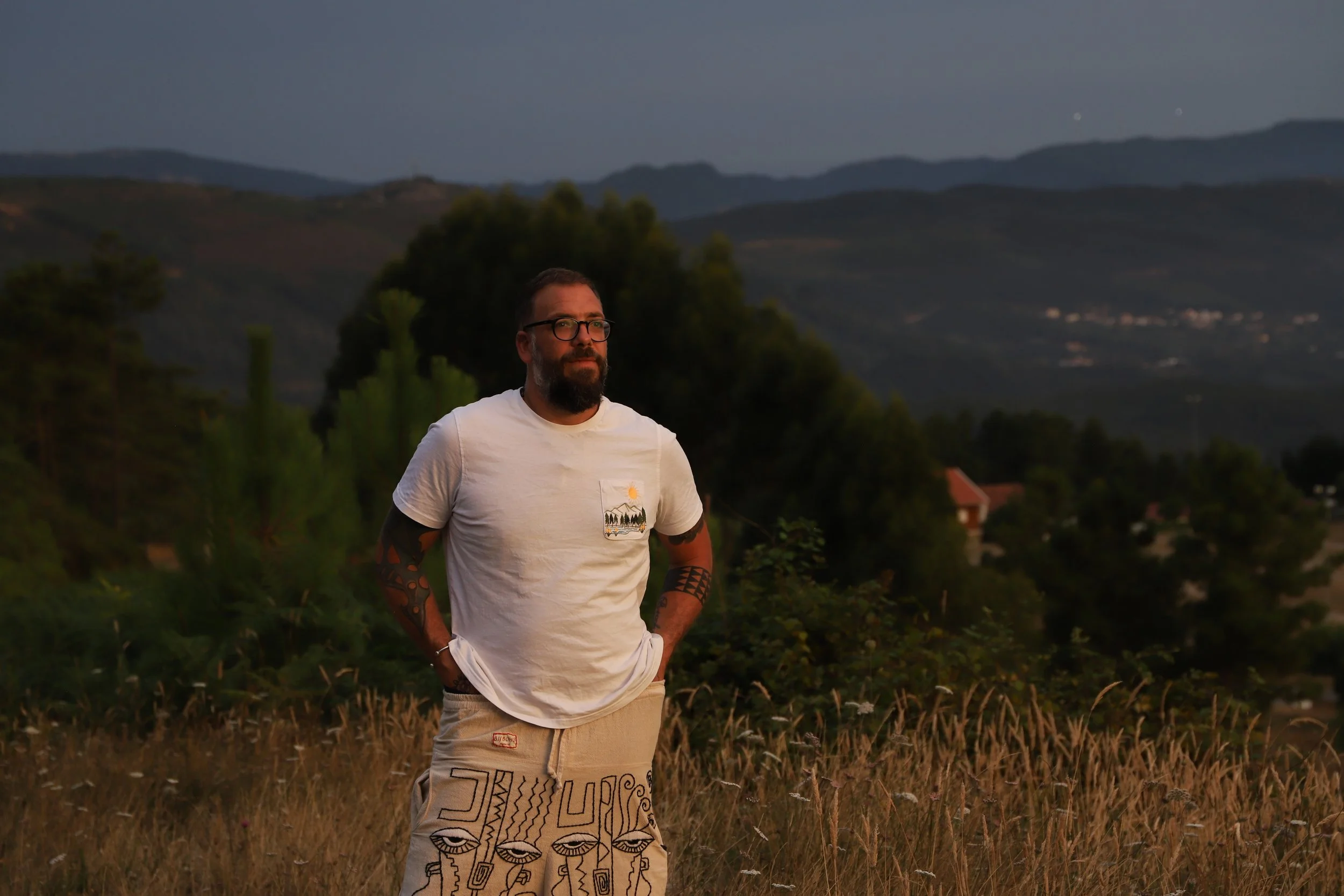 A man with glasses and a beard stands in a field during dusk, wearing a white t-shirt with a small design and beige shorts with patterns and faces, with trees, mountains, and houses in the background.