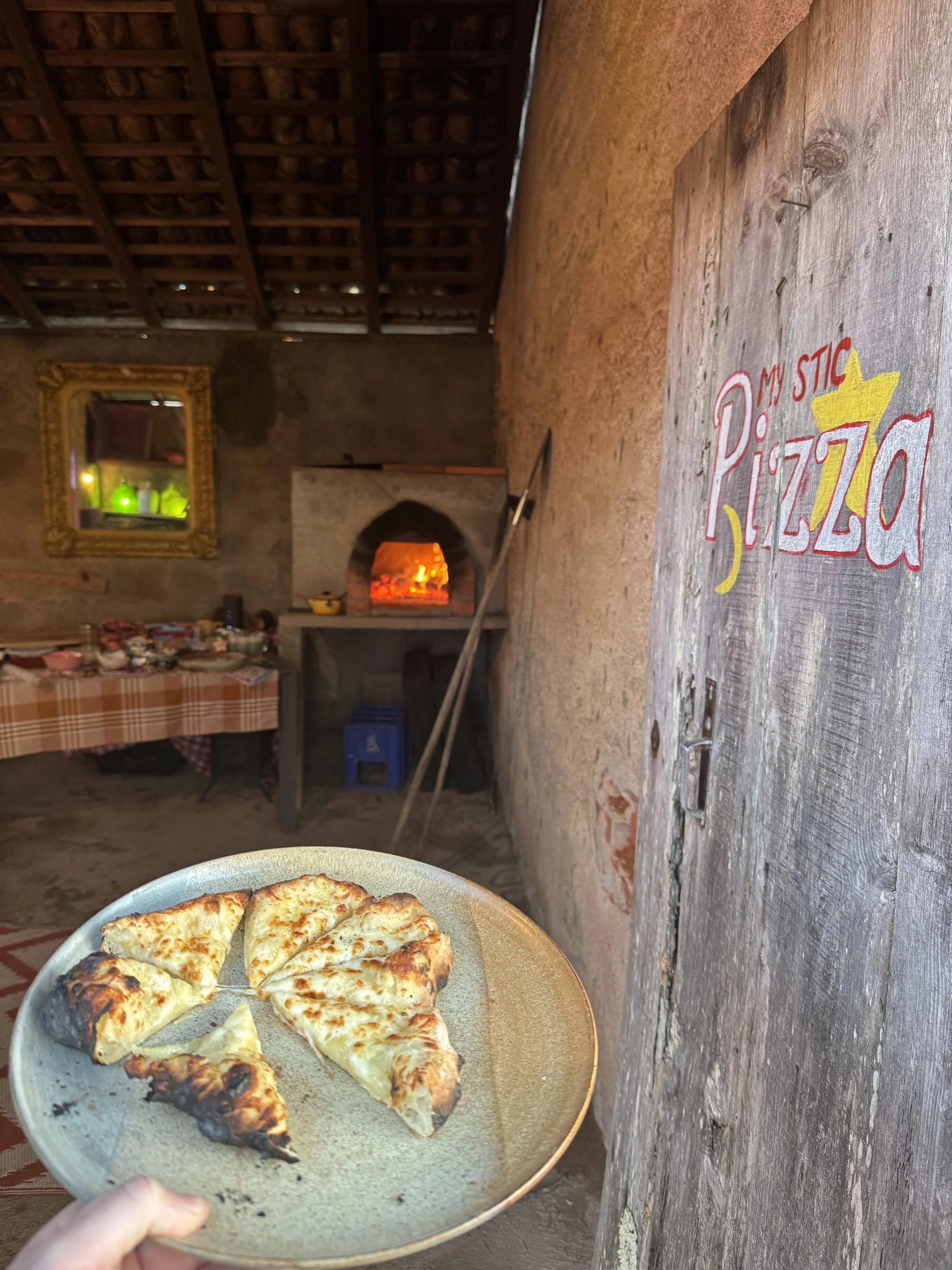A plate of cheese pizza slices being held in front of a rustic indoor setting with a brick oven, a mirror, and a wooden table.
