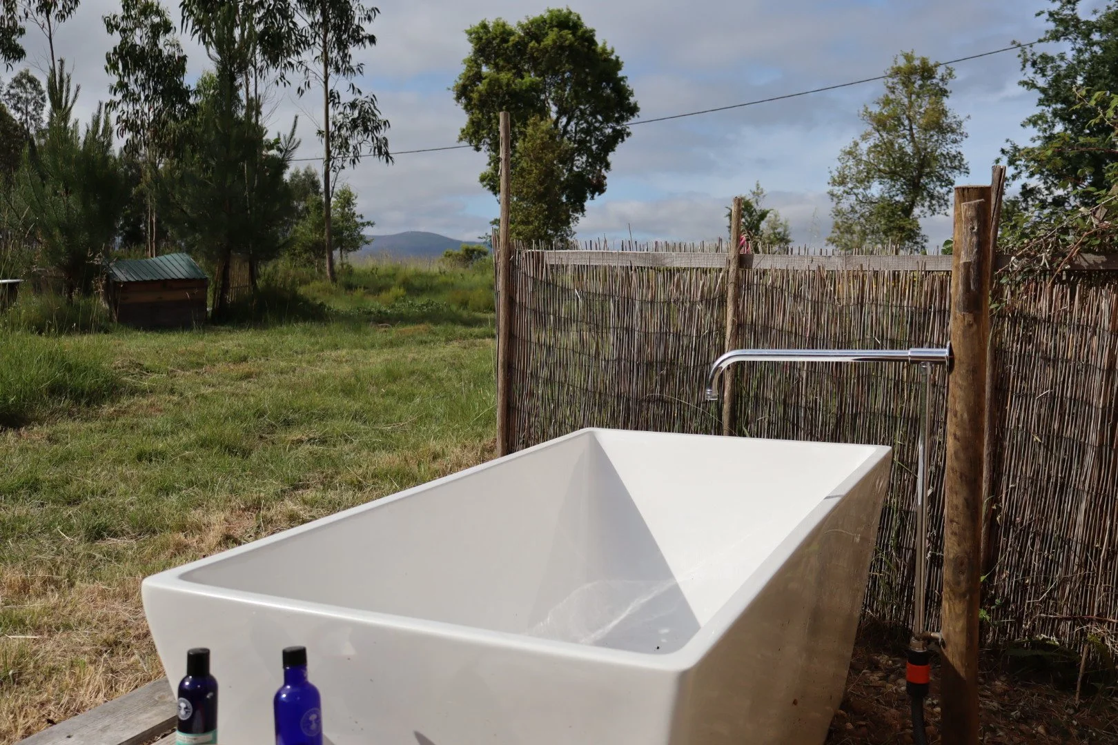 An outdoor scene featuring a white bathtub with black and blue bottles in the foreground, a bamboo fence, and a grassy field with trees in the background under a partly cloudy sky.