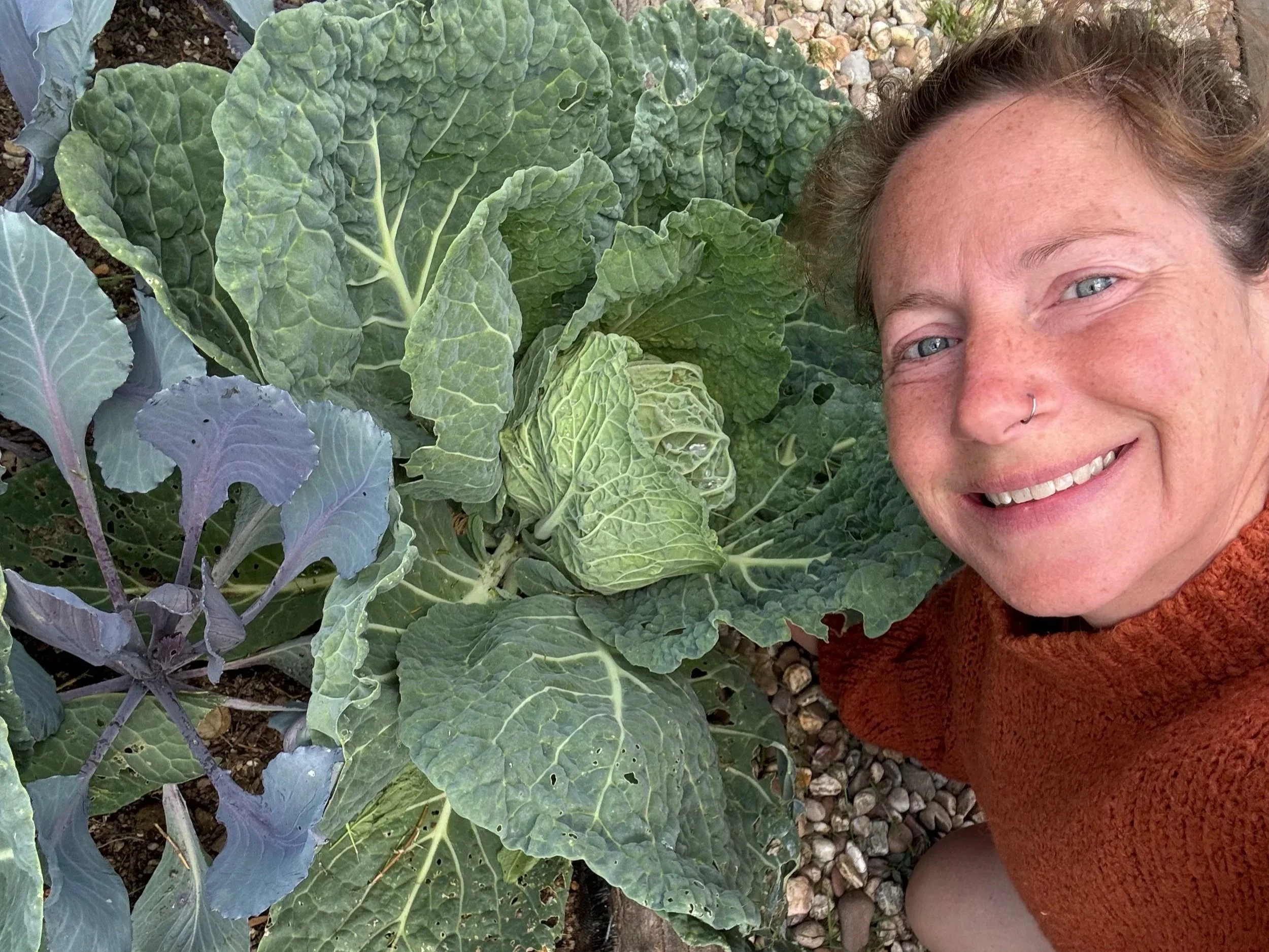 A woman with freckles and a nose ring smiling while lying on the ground next to large green leafy plants in a garden.