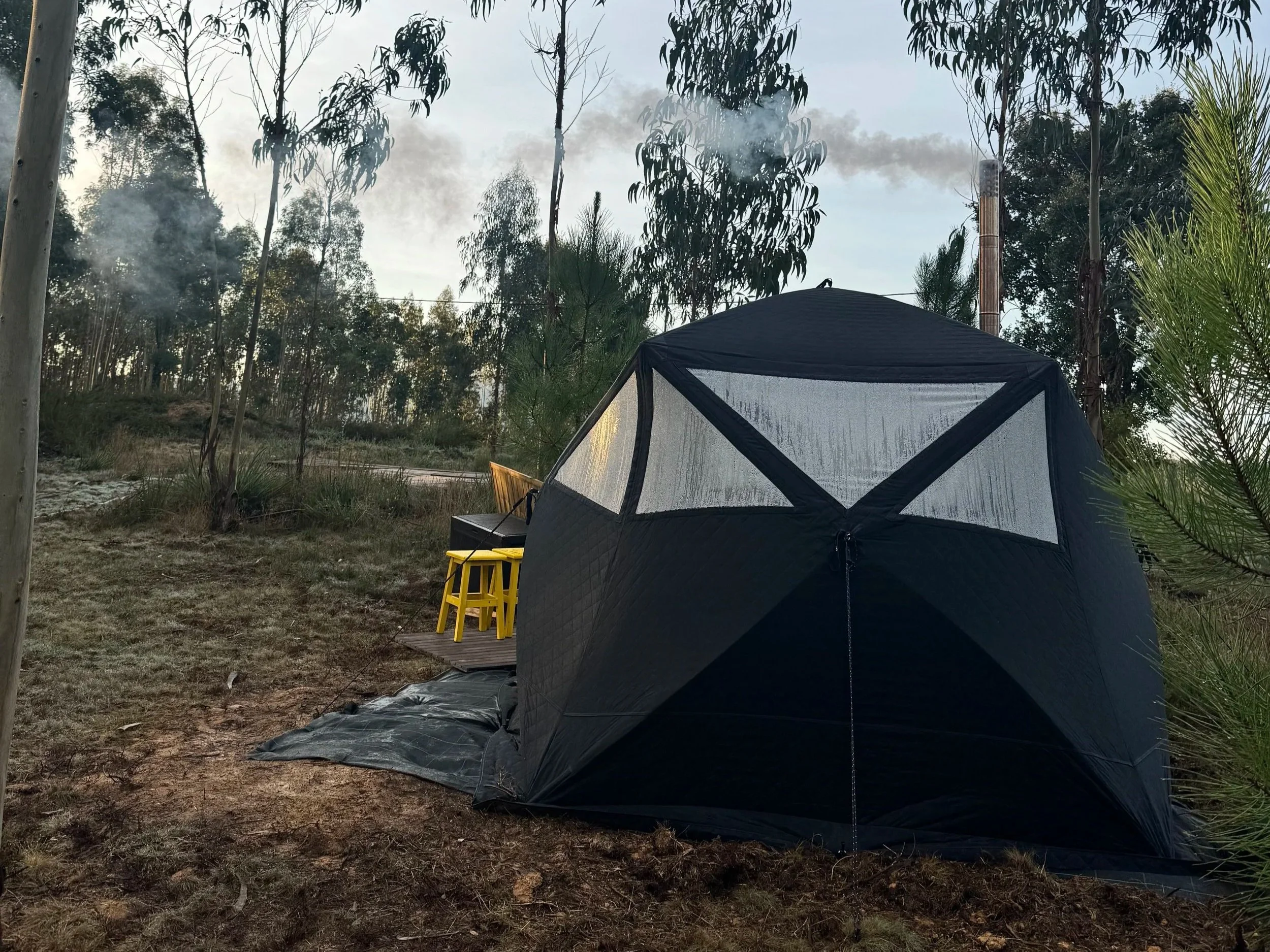A sauna tent on a patch of grass surrounded by trees, with smoke rising in the background and an overcast sky.