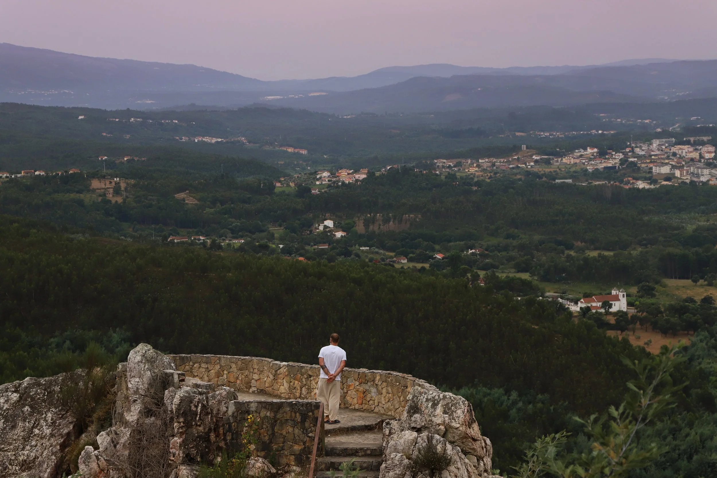 A man standing on a stone wall overlooking a valley with distant mountains, houses, and greenery in the background.