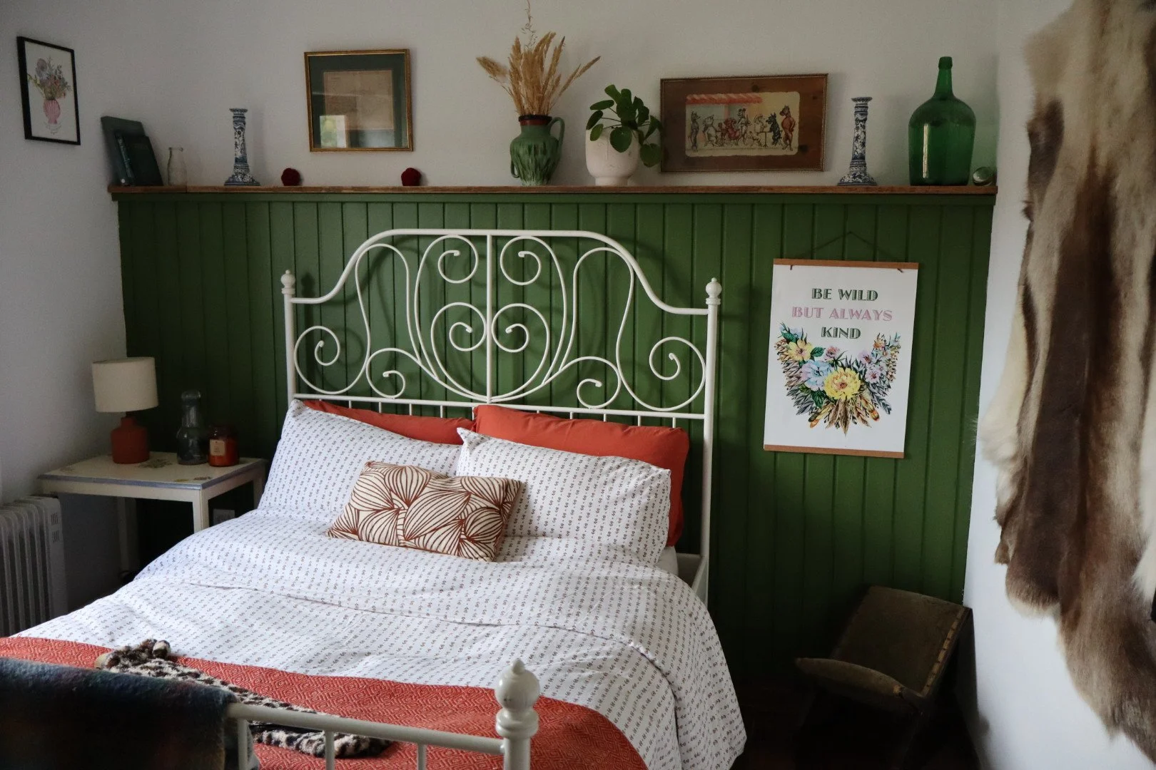 A cozy bedroom with a white metal bed frame, patterned bedding, a small nightstand with a lamp, candles, and decorative items, against a green paneled wall decorated with artwork, a shelf with vases and plants, and a framed sign with floral design and the quote 'Be wild but always kind'.