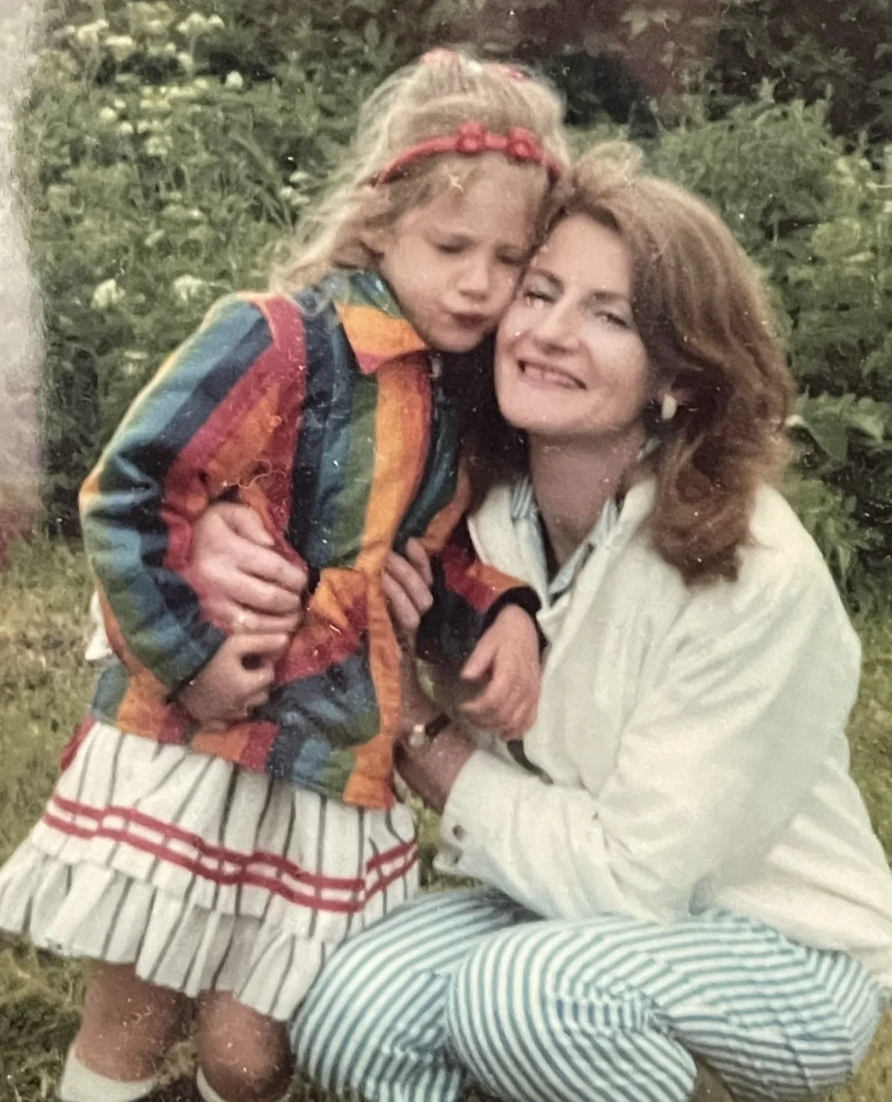 A young girl with long blonde hair and a red headband, wearing a colorful rainbow-patterned jacket and striped skirt, is hugging a woman with curly brown hair, wearing a white jacket, as they sit outdoors in front of greenery.