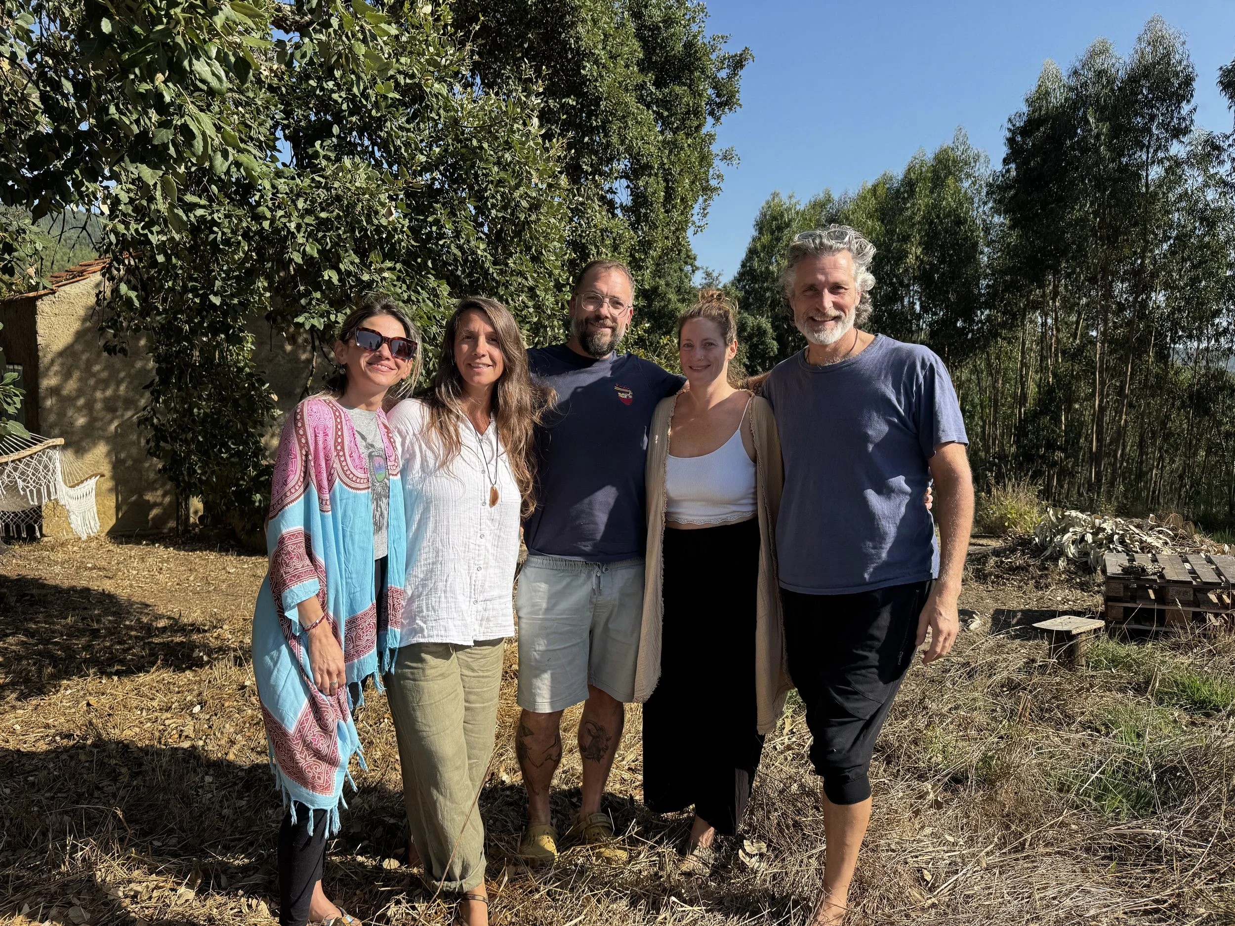 Group of five people standing outdoors under a blue sky, surrounded by trees and dried grass, smiling at the camera.
