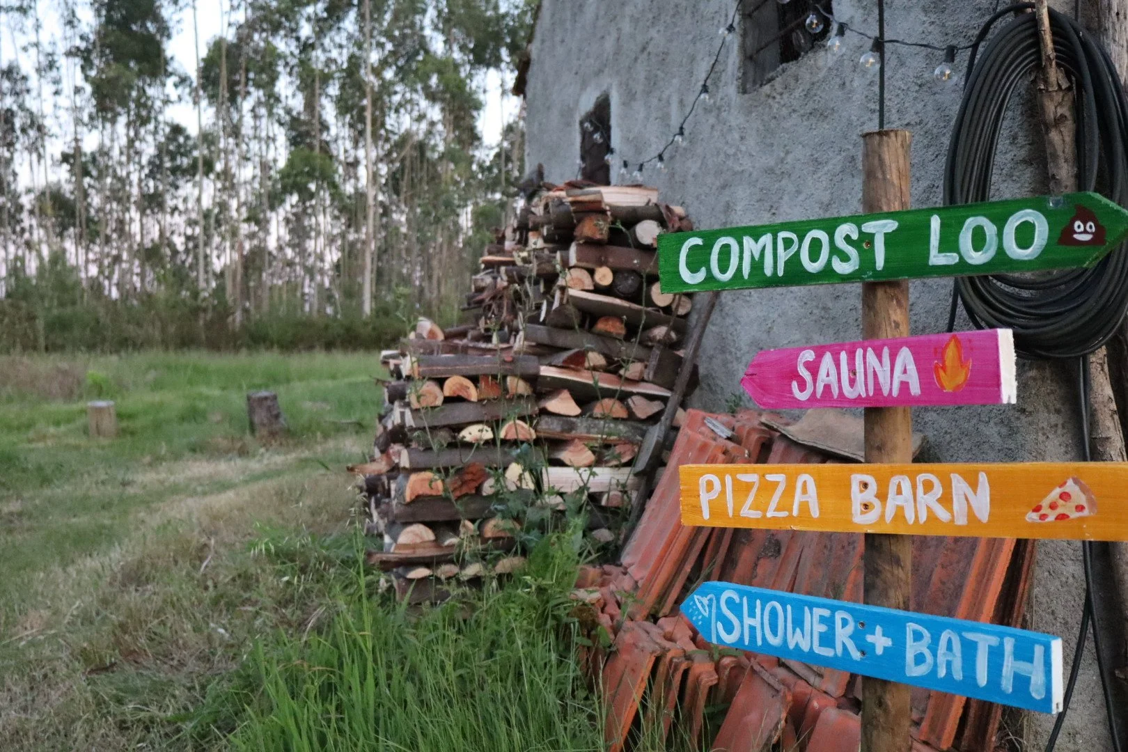 Colorful wooden signs pointing to different outdoor amenities, including compost loo, sauna, pizza barn, and shower and bath, set against a rustic, outdoor setting with a woodpile and trees.