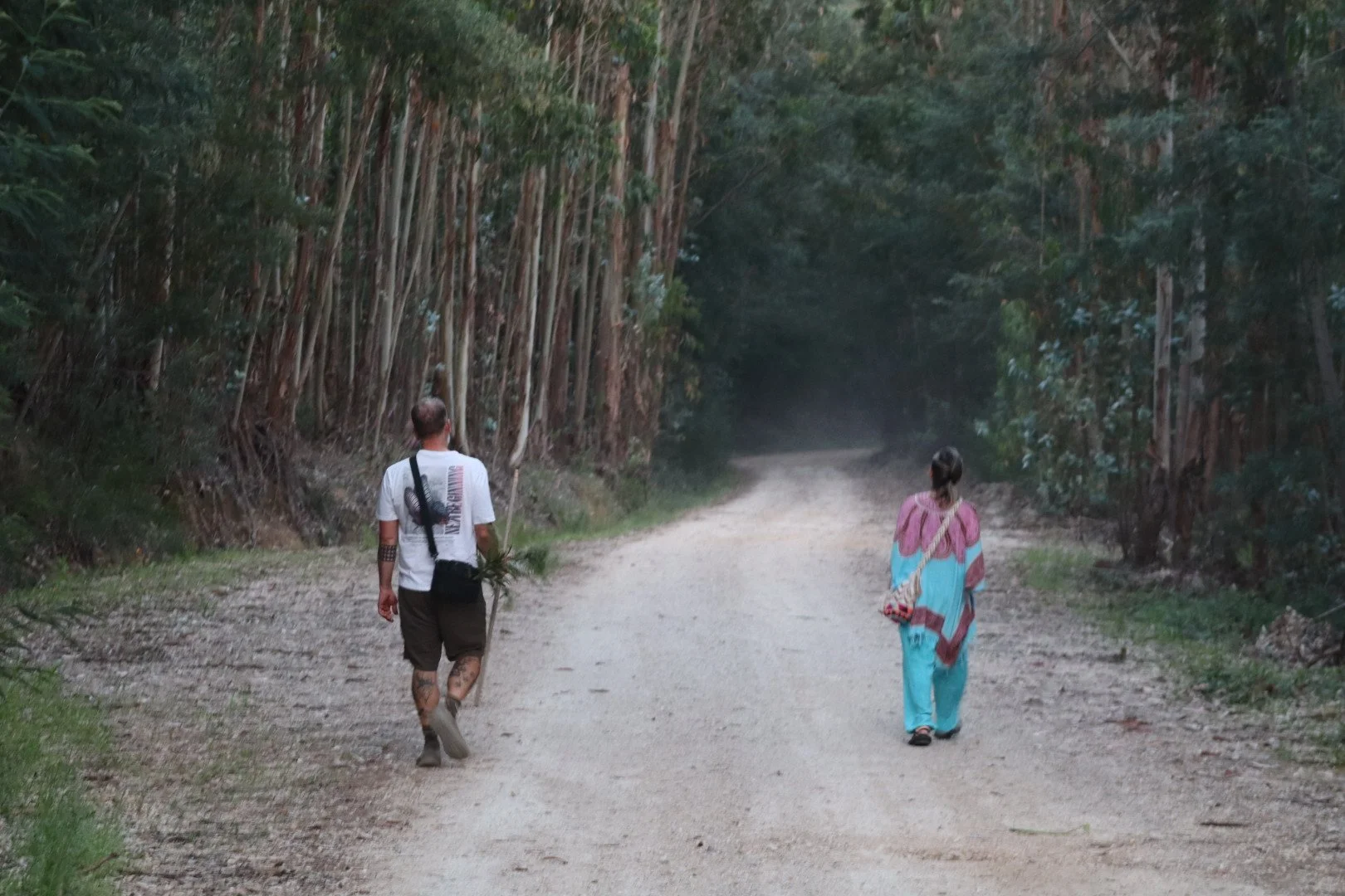 A man and a woman walking on a dirt path through a forested area.