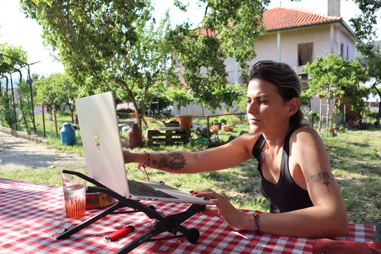 A woman with short dark hair and tattoos on her arms sitting at a red and white checkered table outdoors, reaching toward a small white laptop on a stand, with a glass of water and some snacks nearby, in a backyard with green trees and a house in the background.