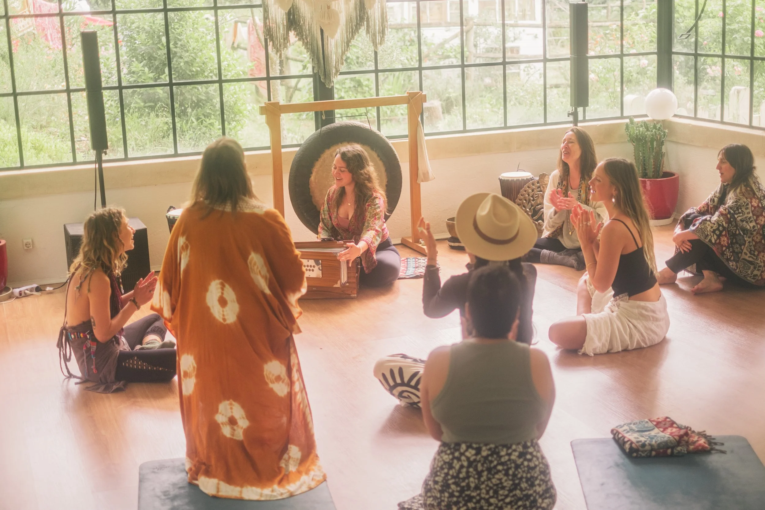 Women and children sitting on the floor in a circle inside a bright room with large windows, participating in a wellness or meditation activity, with a woman playing a musical instrument.