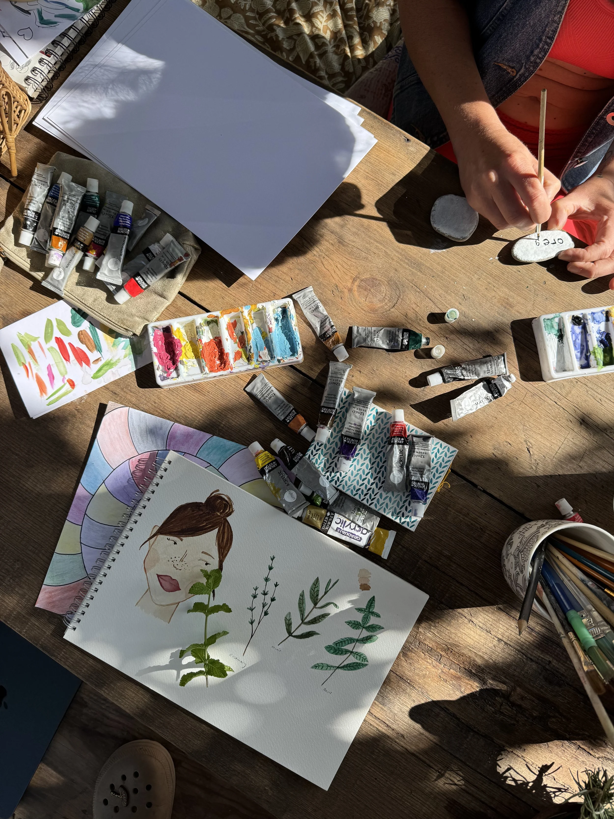 An open watercolor painting of a woman with long hair and freckles, with a green leafy plant on her shoulder, is on a wooden table surrounded by watercolor paints, tubes, brushes, and art supplies. A person's hand is painting on a small stone, with sketchbooks and other art materials nearby.