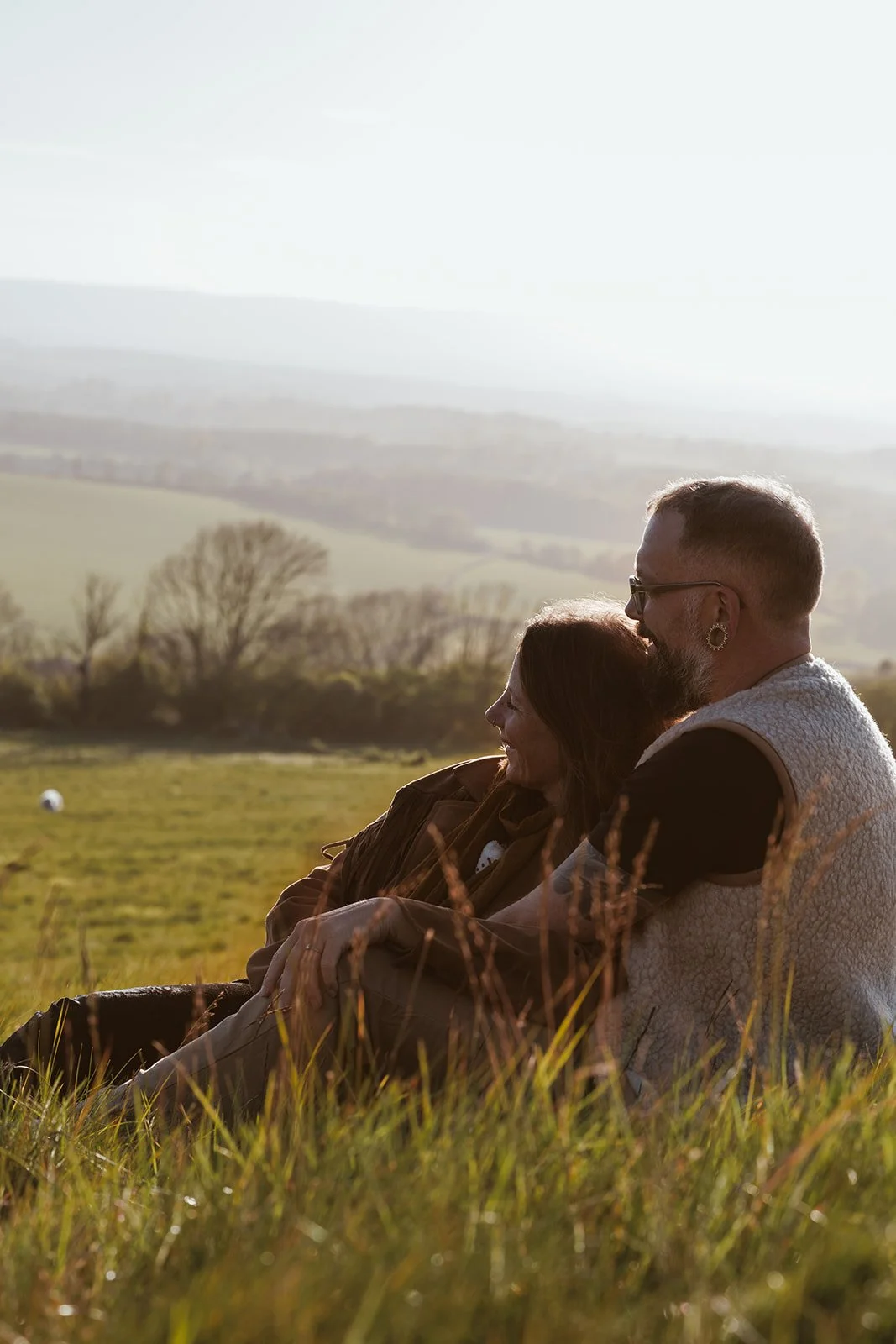 A couple sitting on a grassy field during sunset, with rolling hills and trees in the background, enjoying a peaceful moment together.