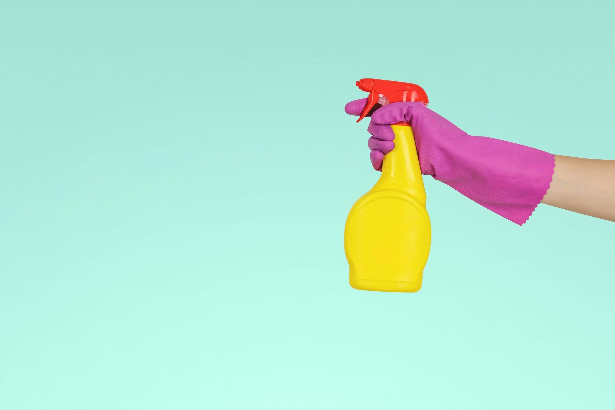 Hand in pinker Gummihandschuh hält eine gelbe Sprühflasche mit rotem Sprühkopf gegen einen hellblauen Hintergrund.