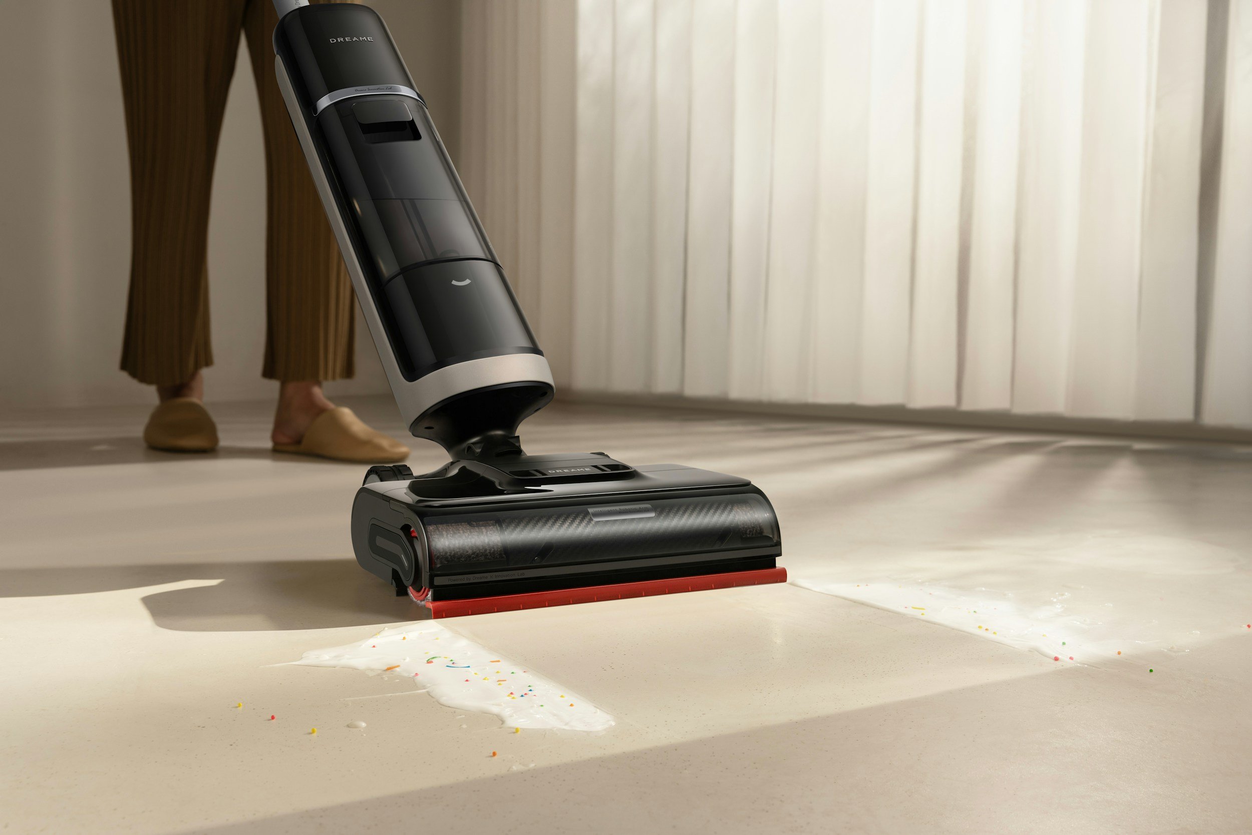 Ein Staubsauger mit einer schwarzen und silbernen Design ist auf einem Boden, der mit bunten Peeps und Süßigkeiten bedeckt ist, während eine Person mit braunen Hosen und beigen Schuhen im Hintergrund steht.