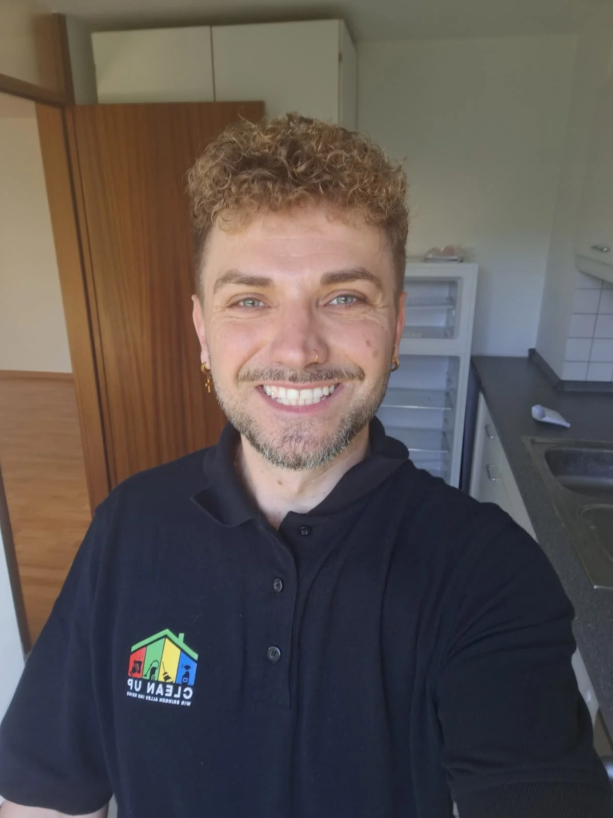 Ein Mann mit lockigem, hellbraunem Haar und Bart, der in einer Küche lächelnd einen Selbstporträt selfie macht.